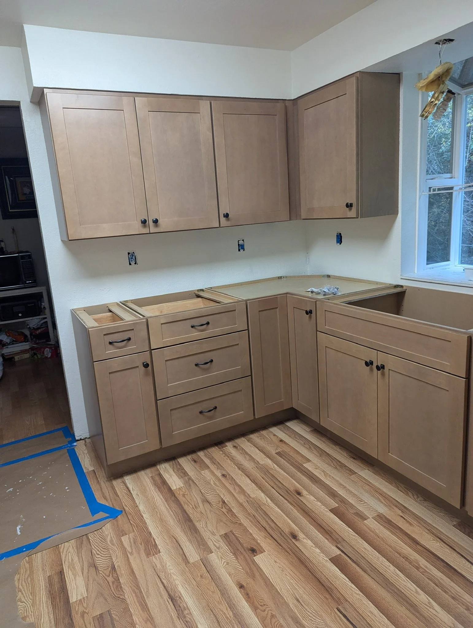 Kitchen cabinets under construction with light wood finish, some drawers and doors installed, open space for appliances, and a window with a view outside.