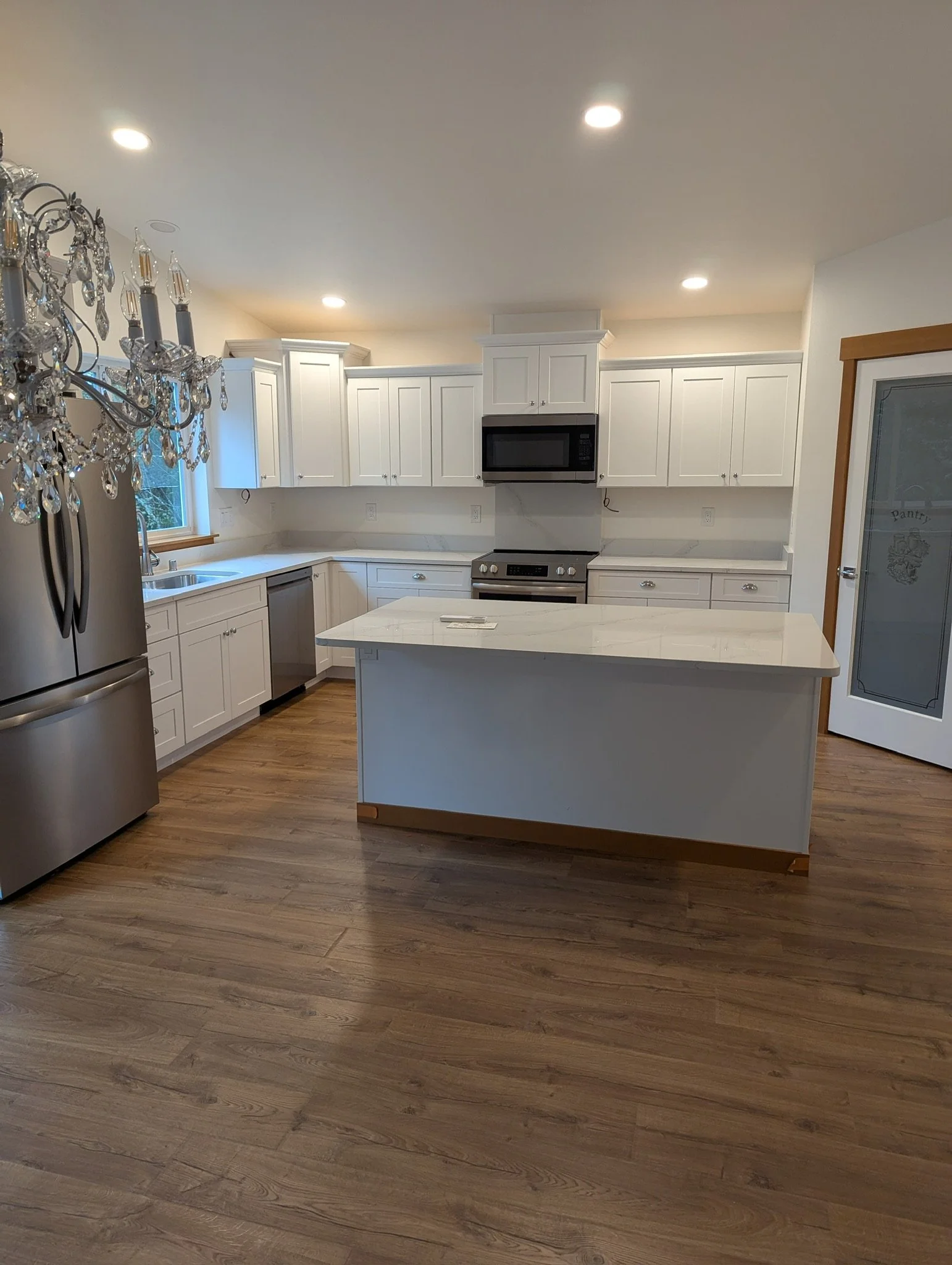 Modern kitchen with white cabinets, marble countertop island, stainless steel appliances, wooden flooring, chandelier, and a pantry door.