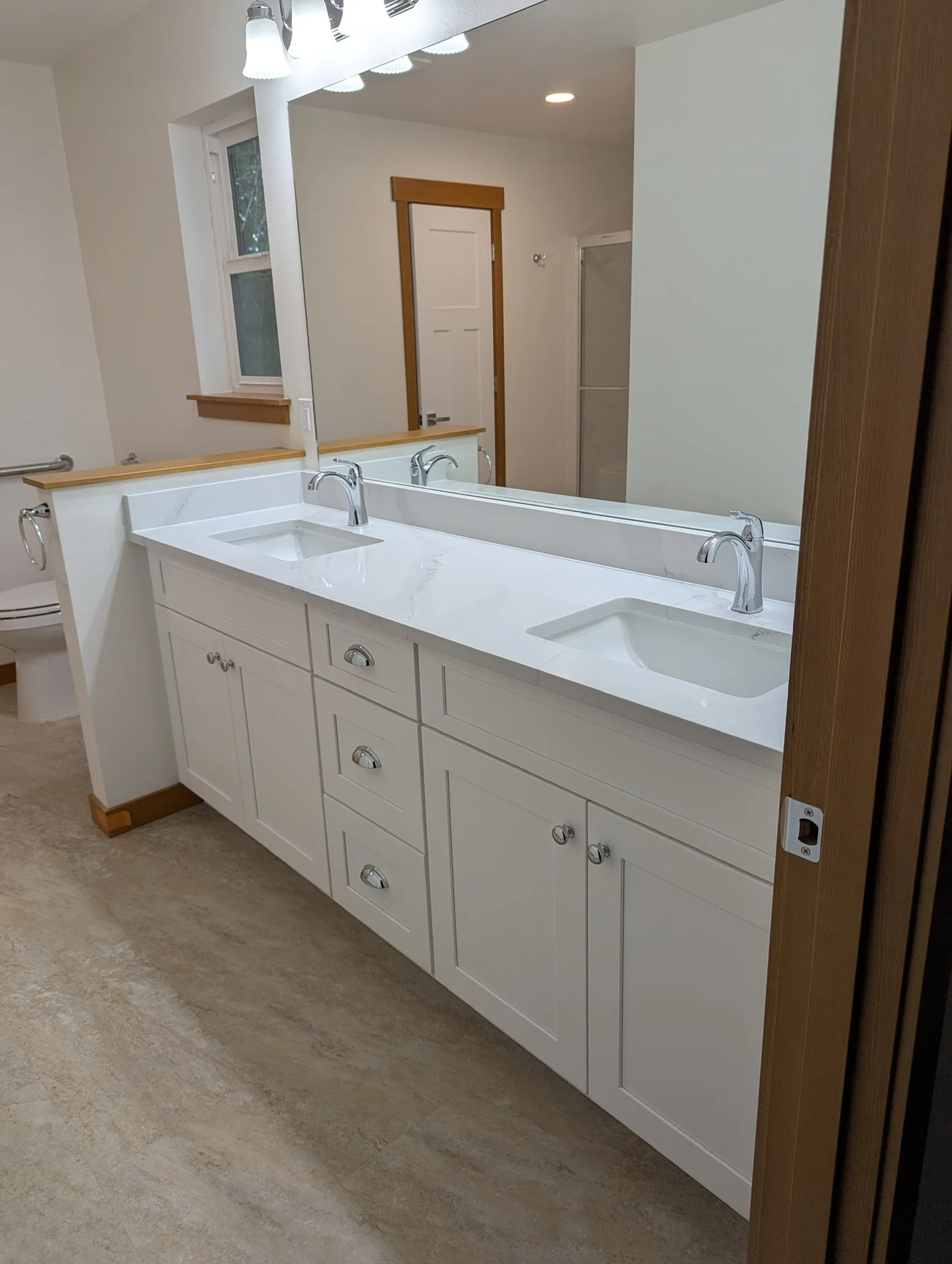 White bathroom vanity with two sinks, a large mirror, and light fixtures above; a window, a toilet, and a shower in the background.