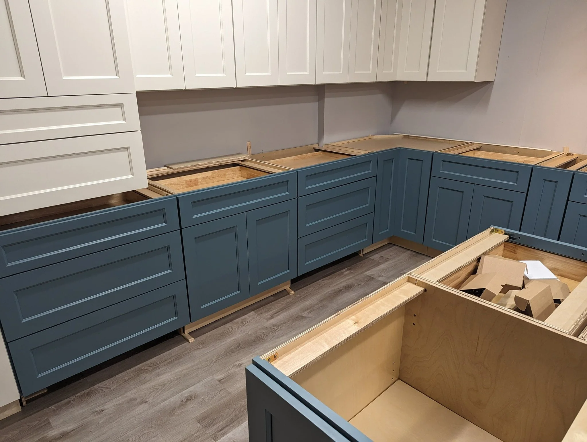 Kitchen cabinets being installed, with blue lower cabinets and white upper cabinets, and some cabinets still unfinished or in construction.