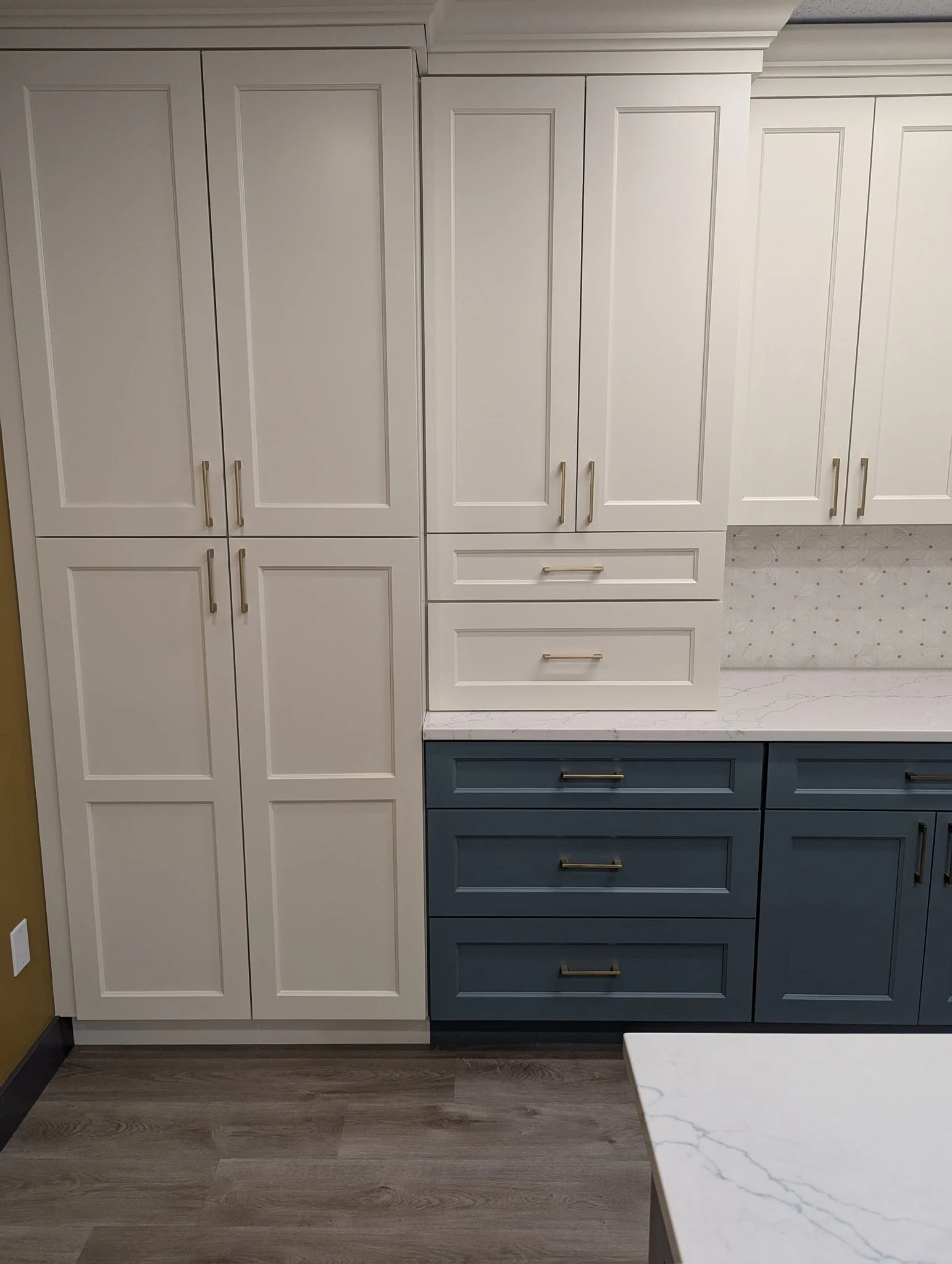 White and blue kitchen cabinets with gold handles, white marble countertop, and wood laminate flooring.