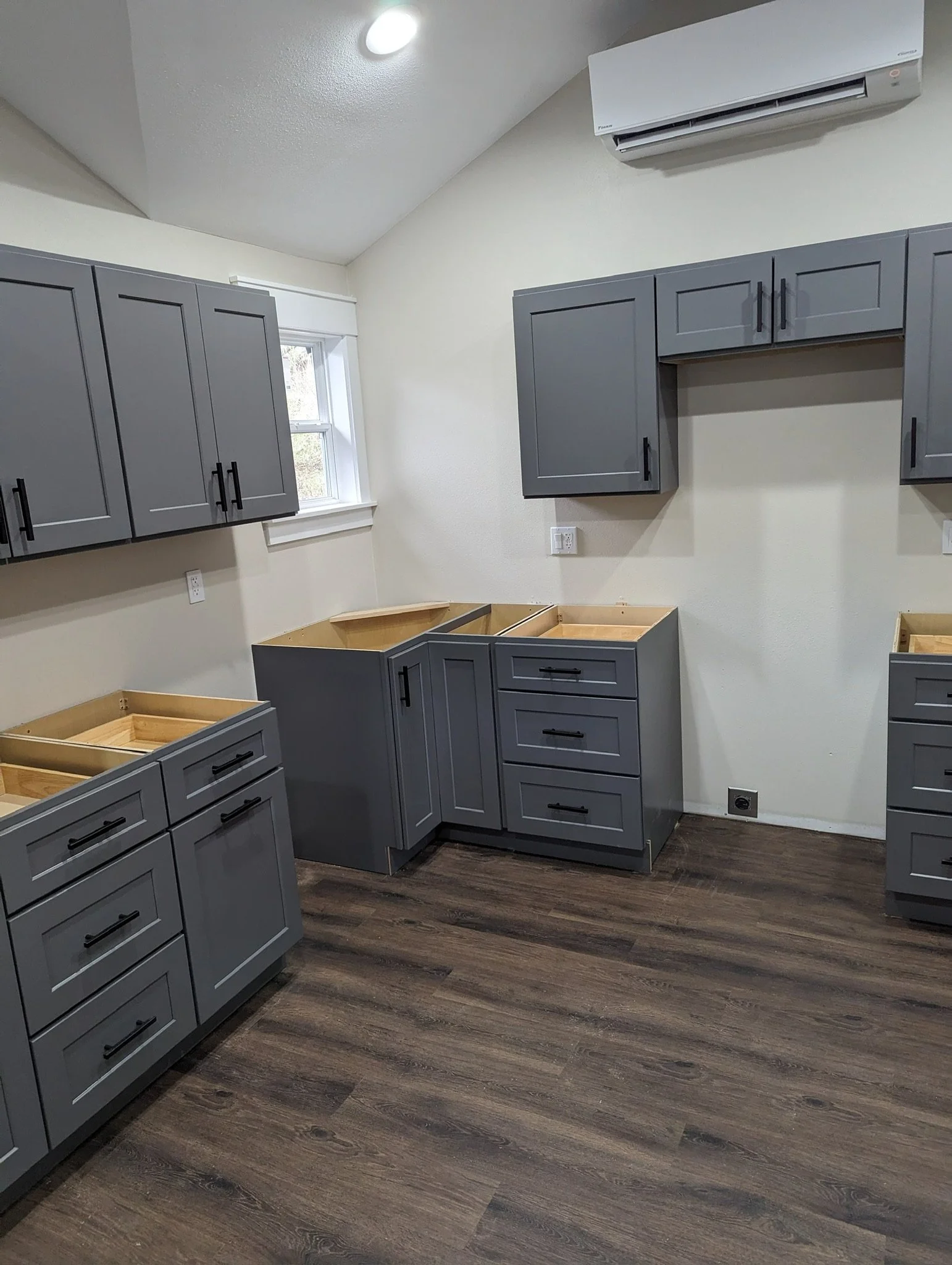 Kitchen with gray cabinets, some without countertops, and a wood floor. There is a window on the wall, with an air conditioning unit above.