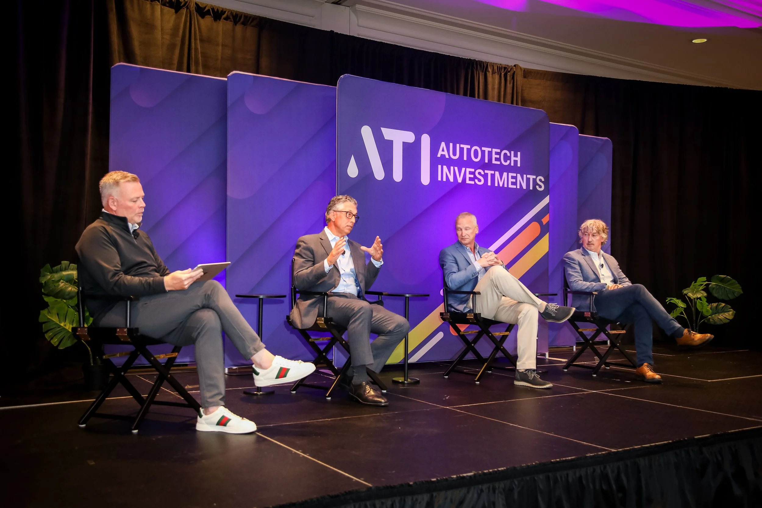 Four men seated on a stage in chairs, participating in a panel discussion at Autotech Investments, with a large purple and blue backdrop featuring the event's logo and name.