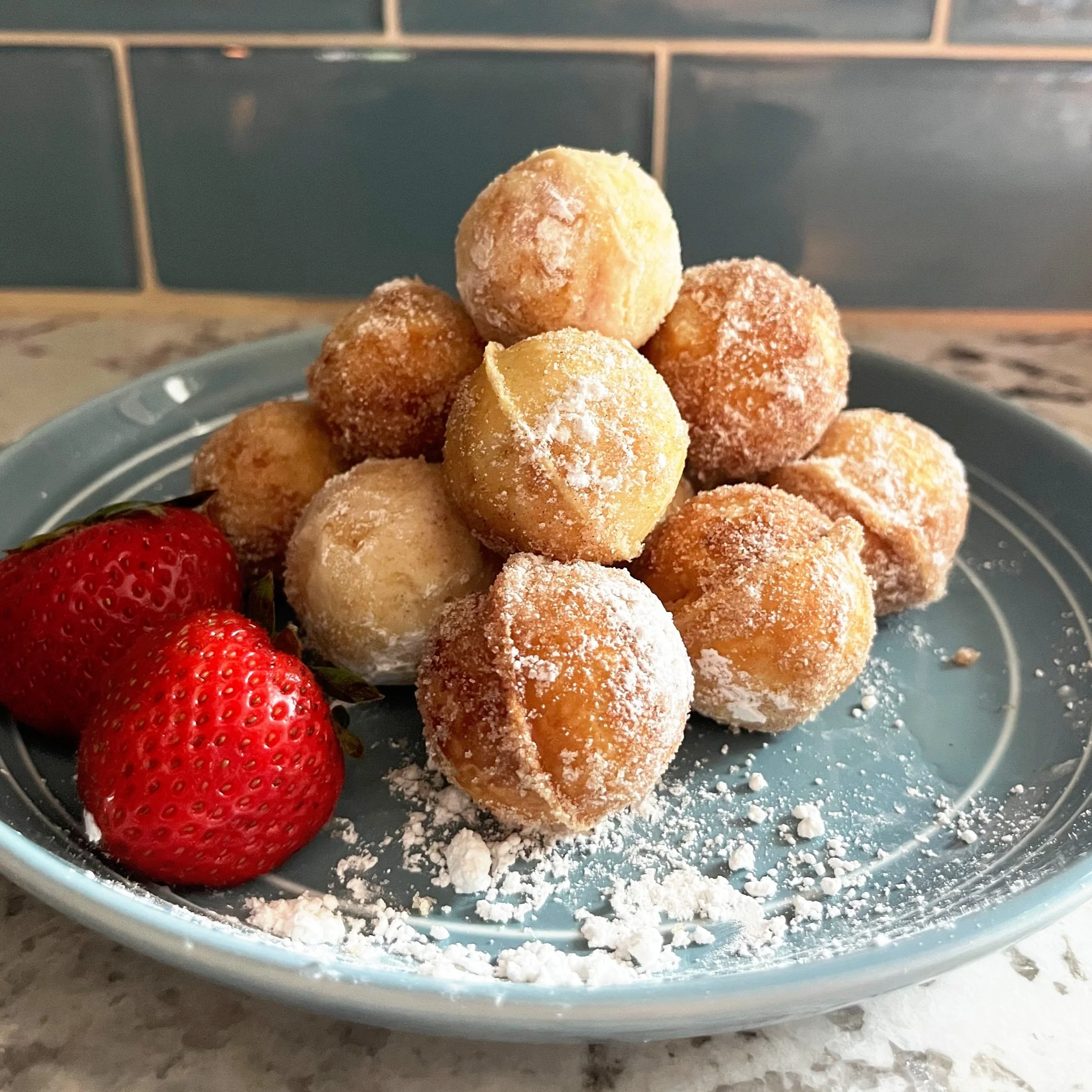 A plate of powdered sugar-covered mini doughnuts with two strawberries on the side.