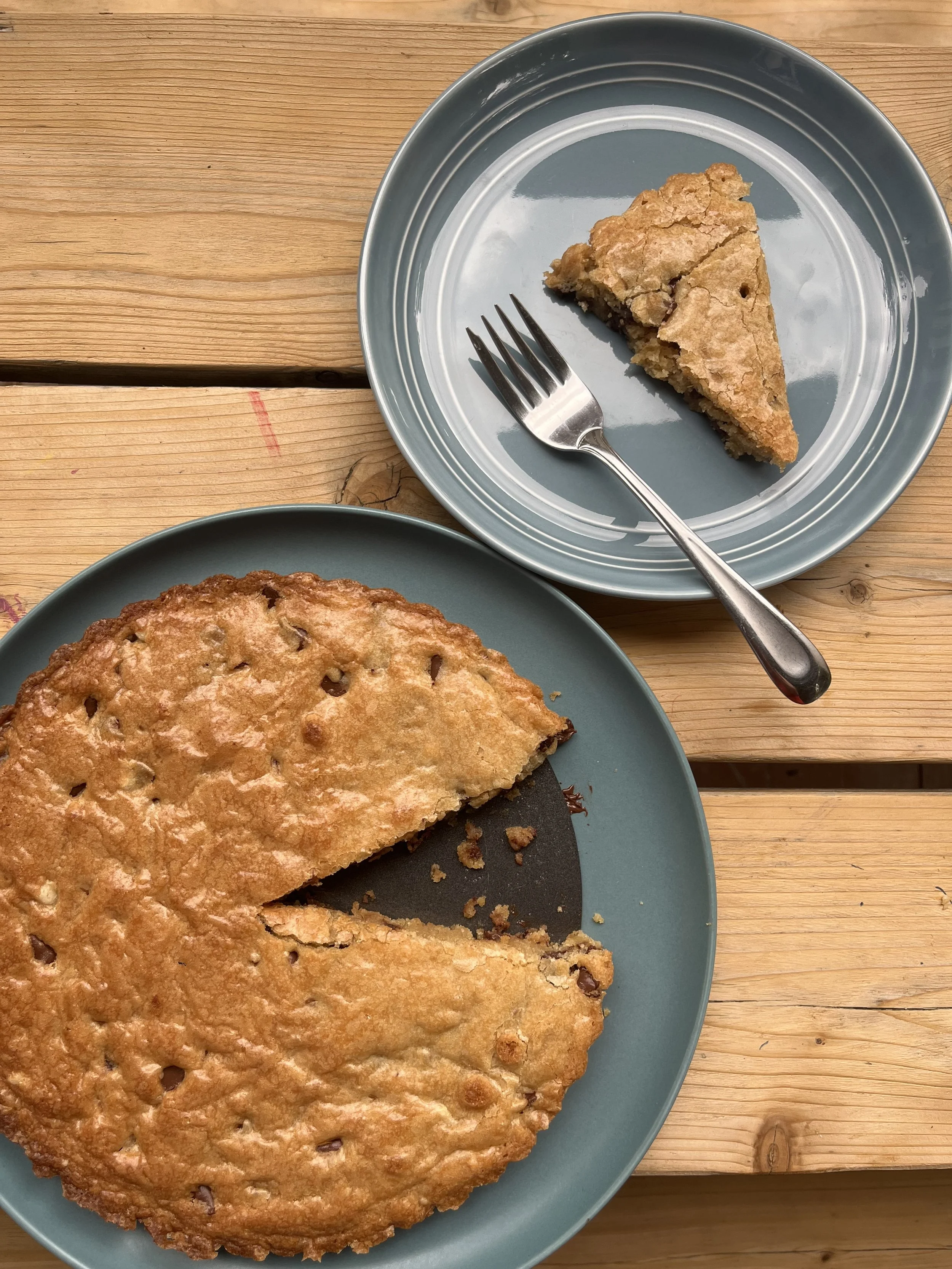 A partially eaten chocolate chip cookie pie on a dark teal plate. A slice of the pie is on a matching plate with a fork, placed on a light wooden table.