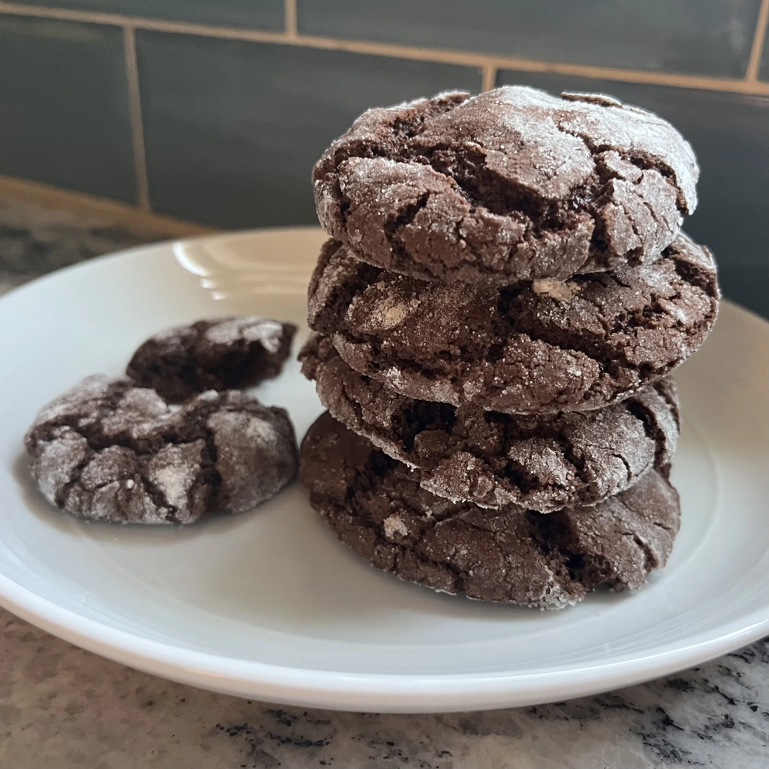 Stack of four chocolate cookies with sugar crystals on a white plate, with some broken pieces nearby.