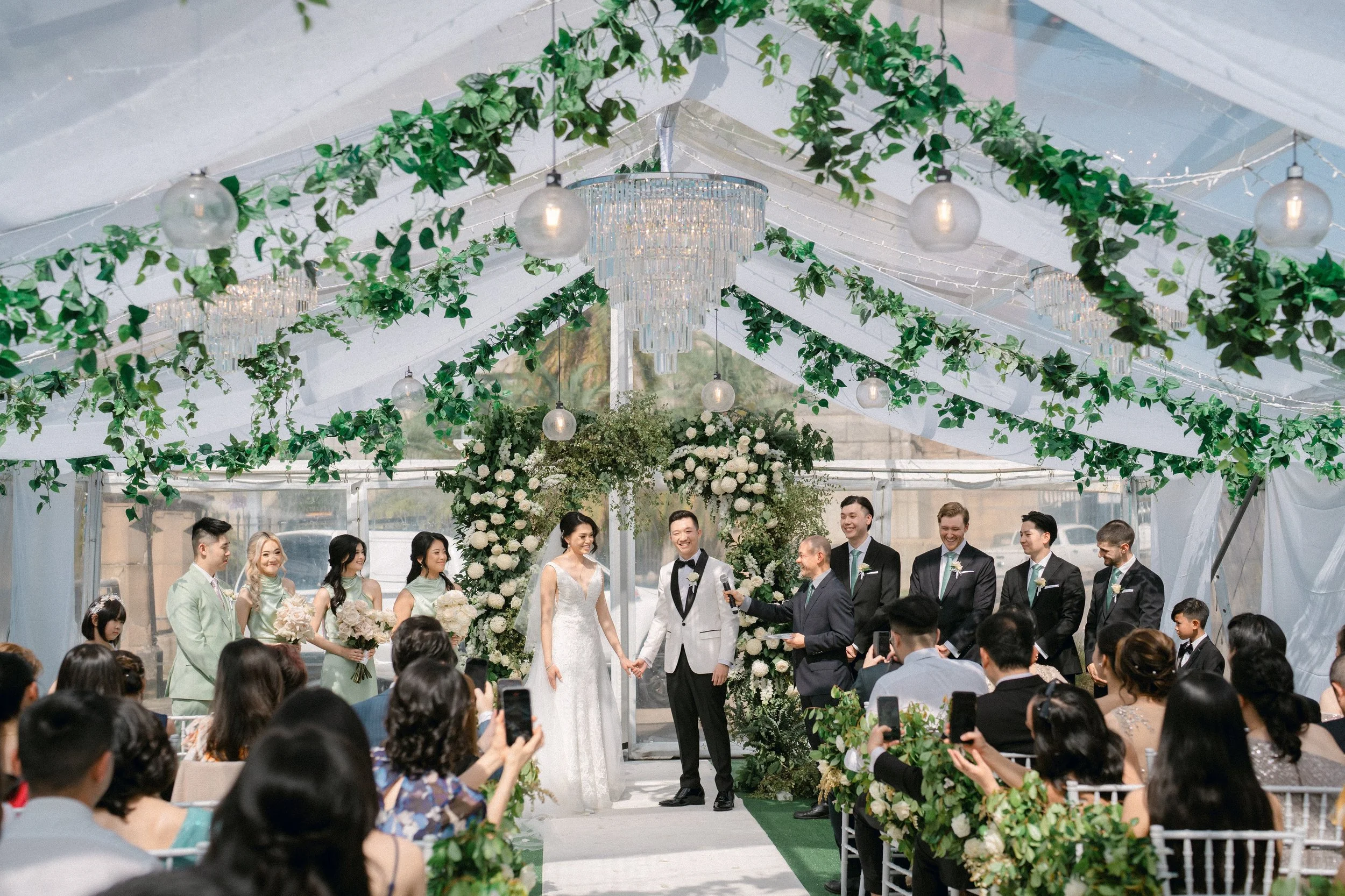 Wedding ceremony under glass marquee with lush greenery ceiling installation