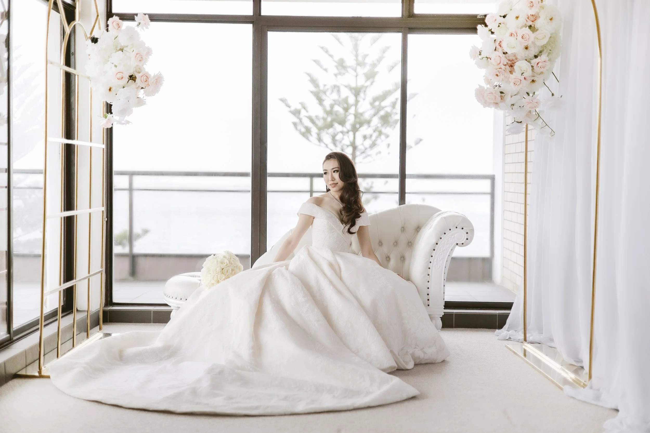 Bride in her white gown on a luxury white lounge with gold frames and flowers in the backdrop