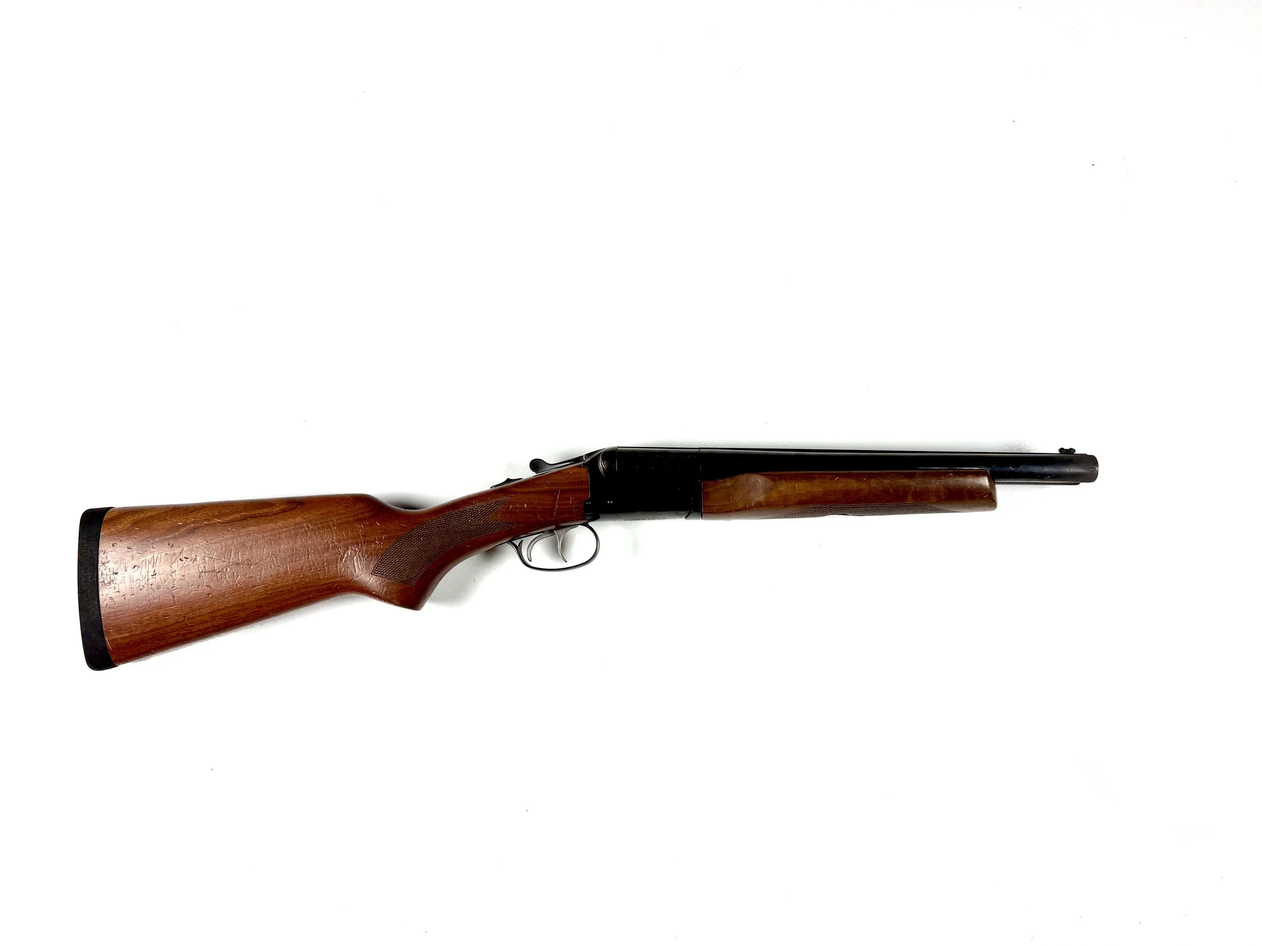 A break-action shotgun with a wooden stock and black barrel, lying horizontally on a plain white background.
