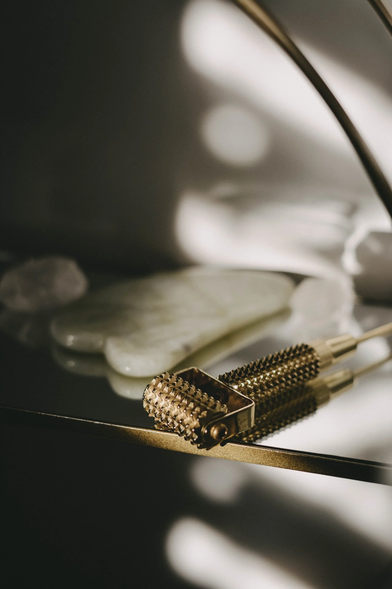 Close-up of a textured gold facial roller and jade scraping tool on a reflective surface with soft lighting.