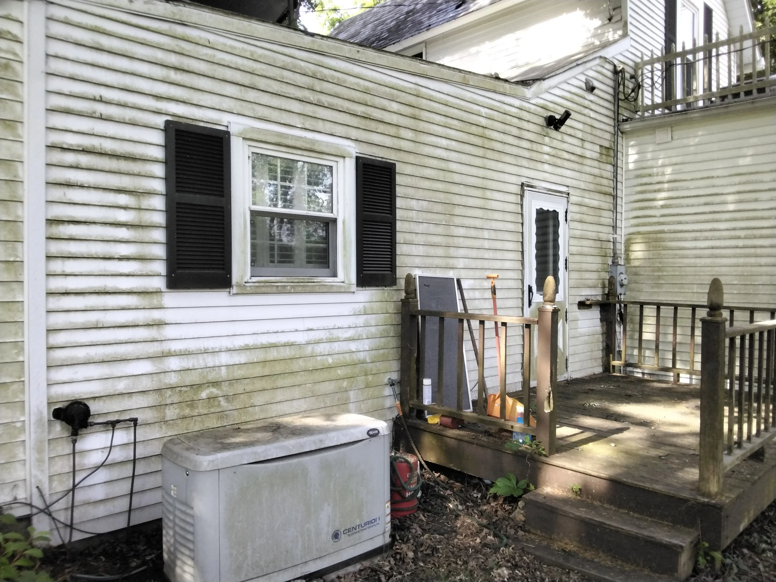 Back porch of a house with white siding and black shutters, showing a window, a door, and a small wooden deck with tools and equipment, including a box fan, a vent pipe, and an outdoor air conditioning unit.