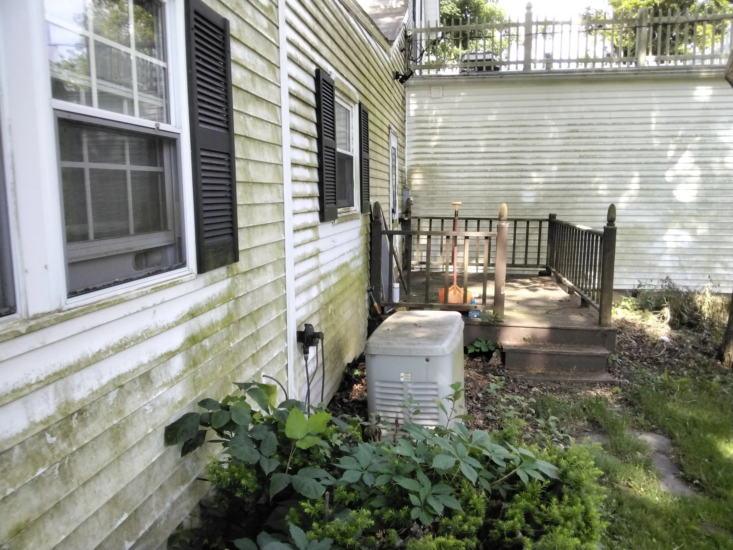 Backyard with weathered siding, three windows with black shutters, a small wooden deck with railings, stairs, gardening tools, and overgrown plants.