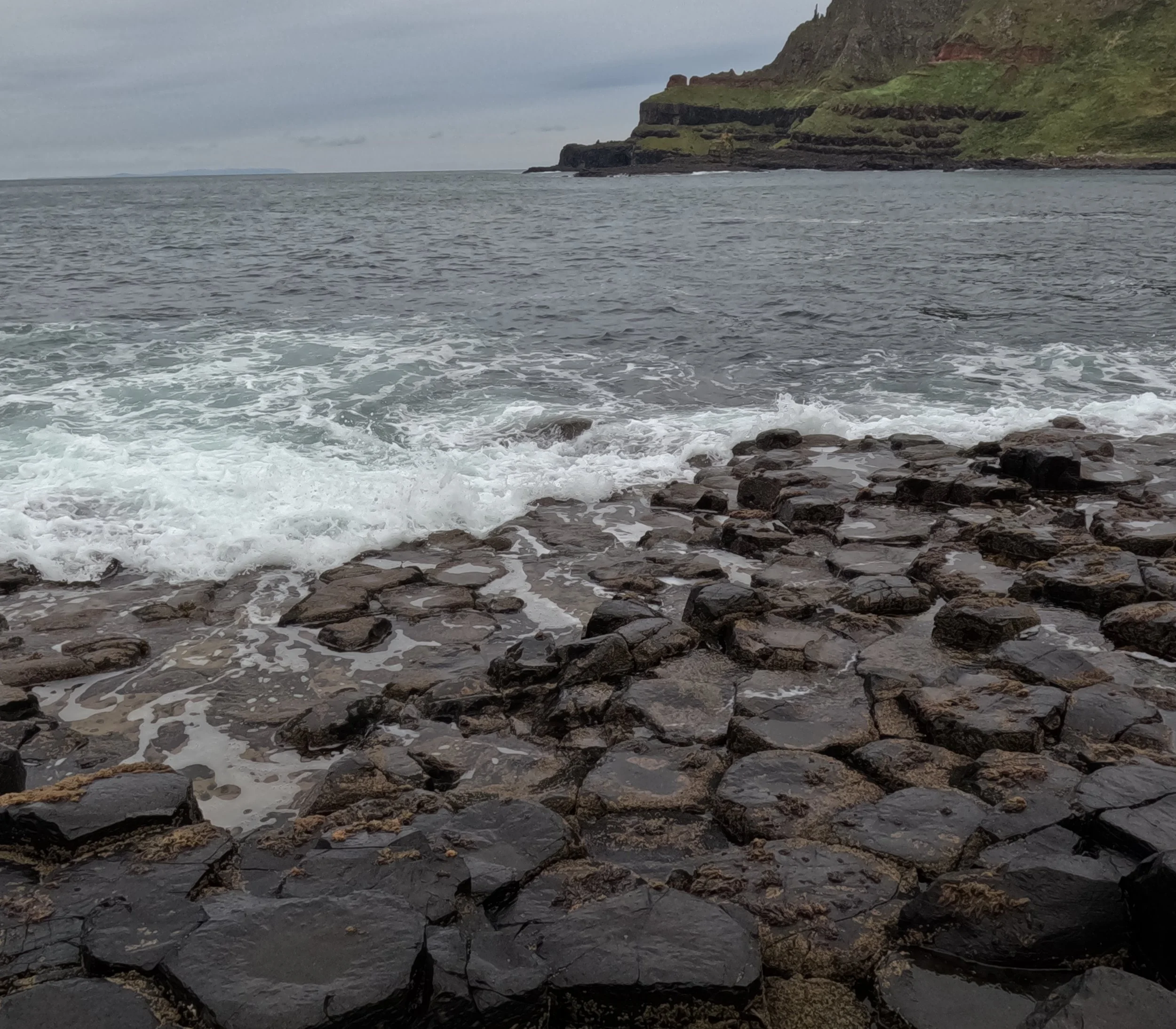 Rocky shoreline with waves crashing against dark rocks, and a green cliff in the background near a cloudy sky.