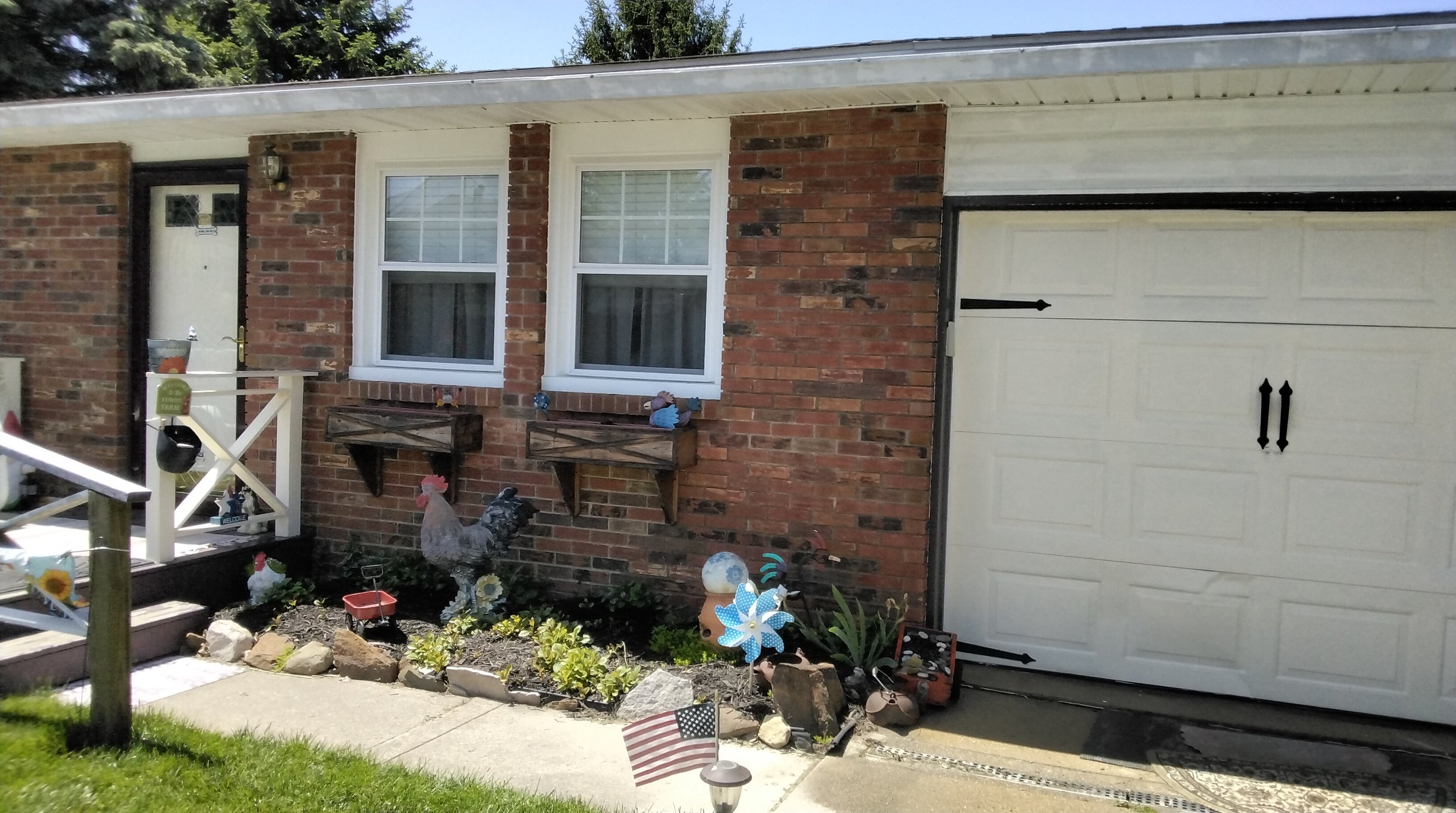Front of brick house with white garage door, two windows, and decorated garden with rooster figure, windmill, small American flag, and various garden ornaments.
