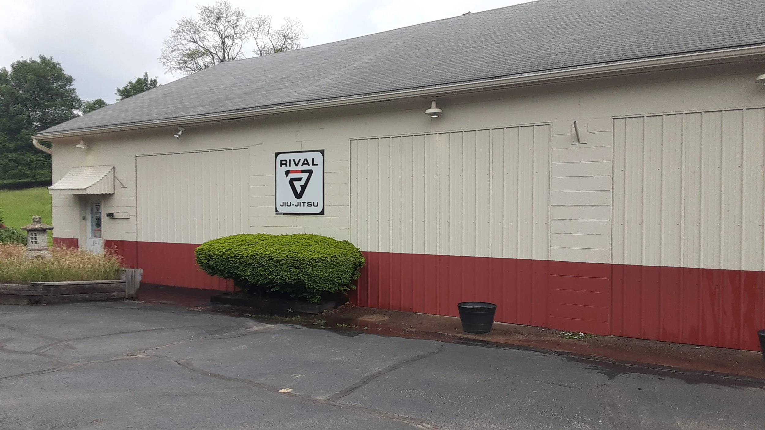 Building with a sign for Rival Jiu-Jitsu, featuring a white exterior with a red lower section, a small awning over a side door, a bush in front, and two black pots on the ground.