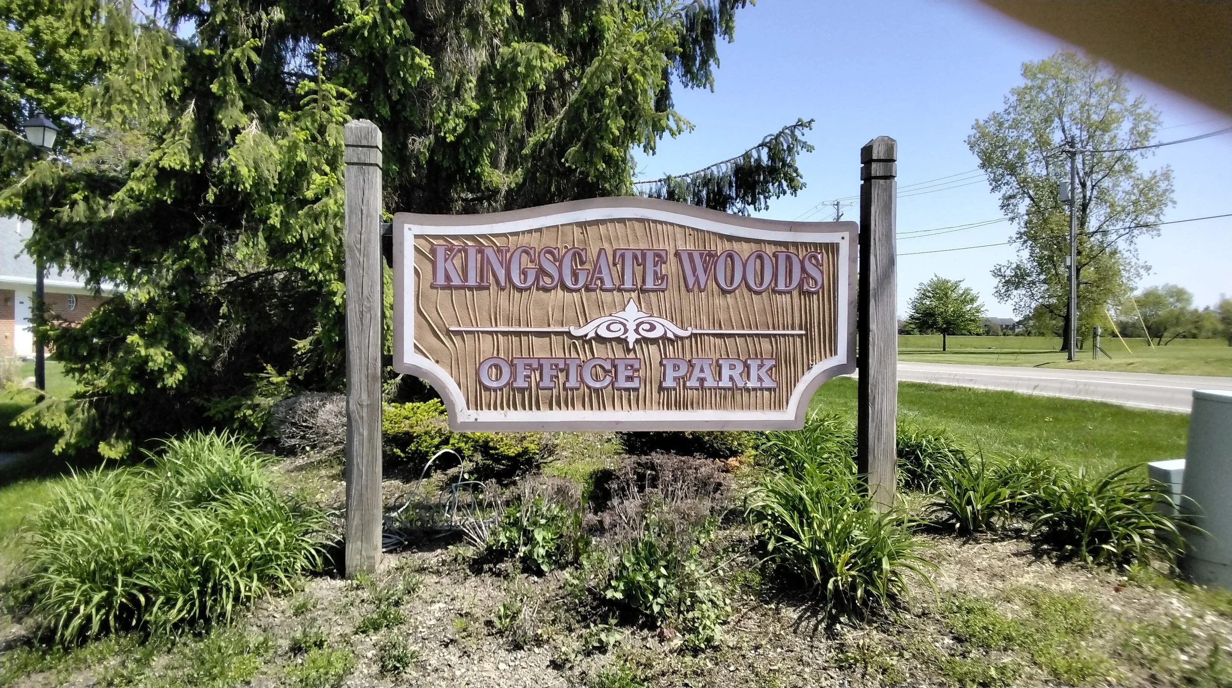 A sign with wooden texture that reads 'Kingsgate Woods Office Park' surrounded by greenery and trees.