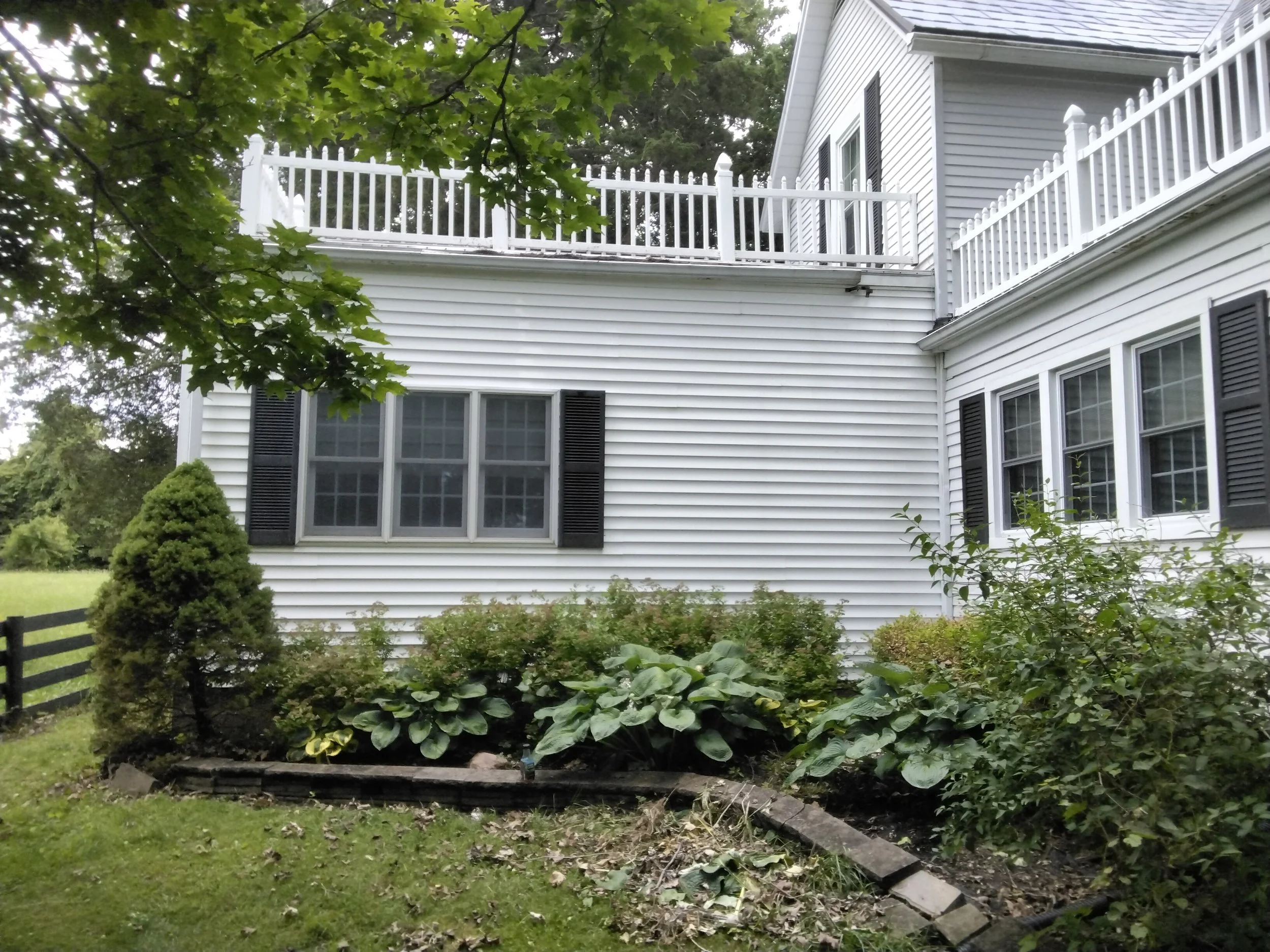 White house with black shutters, multiple windows, and a second-story balcony with white railing. Green bushes and a small garden in the front yard, with a tree partially visible in the top left corner.