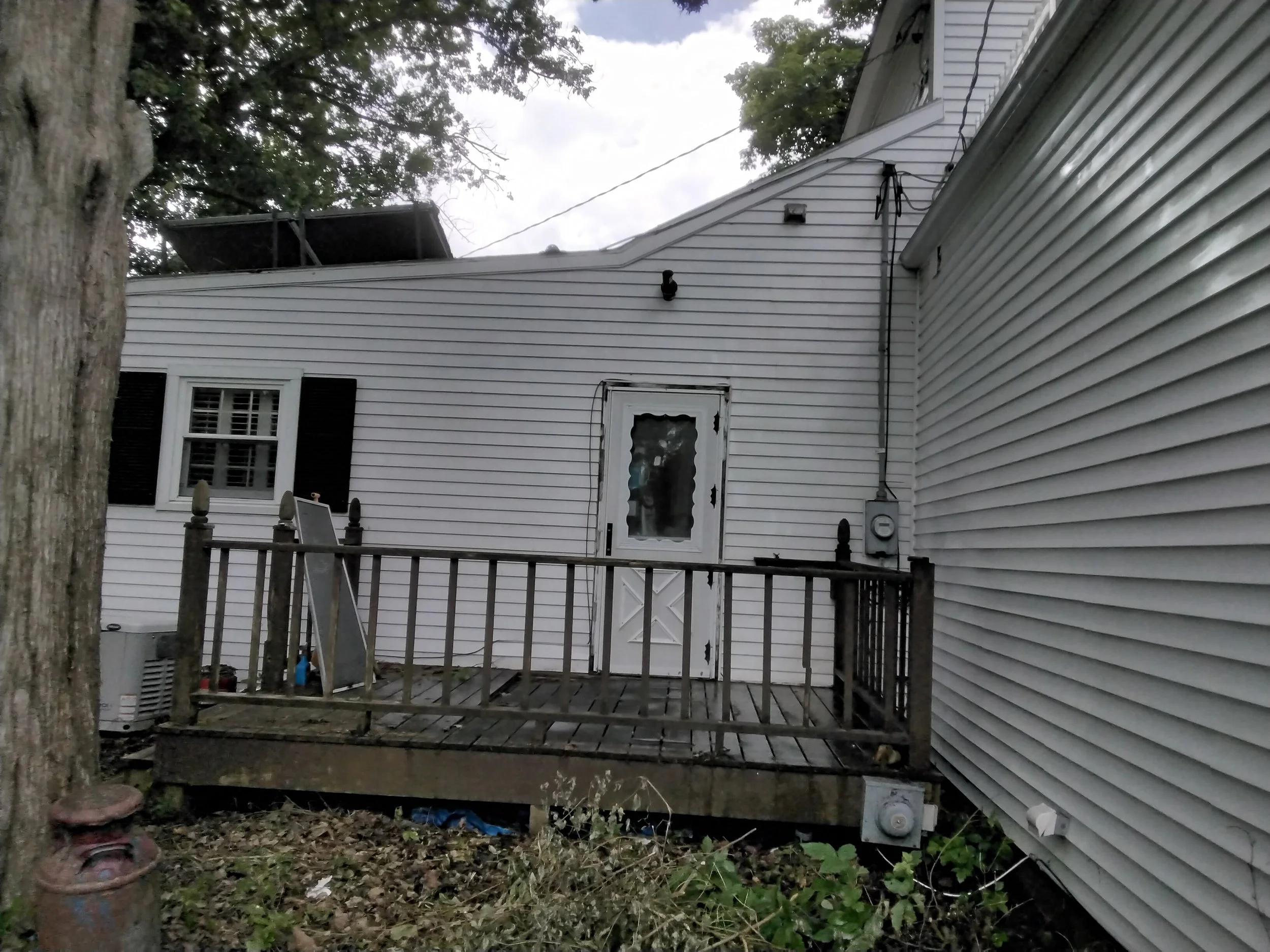 Backyard deck with white house siding, a door, a window, black shutters, and a large tree in front.