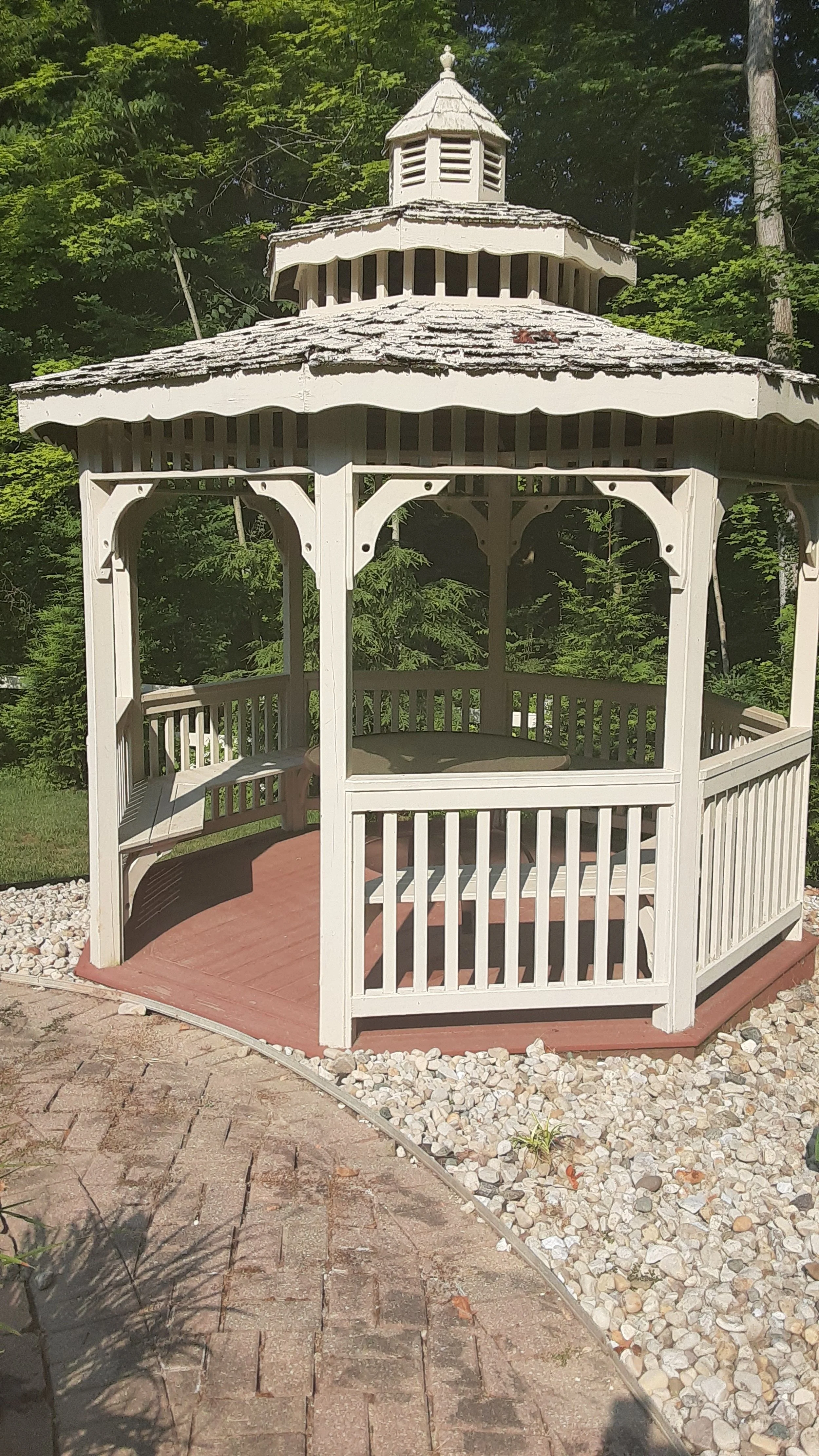 A white wooden gazebo with a shingled roof and small cupola, surrounded by trees and a landscaped yard.