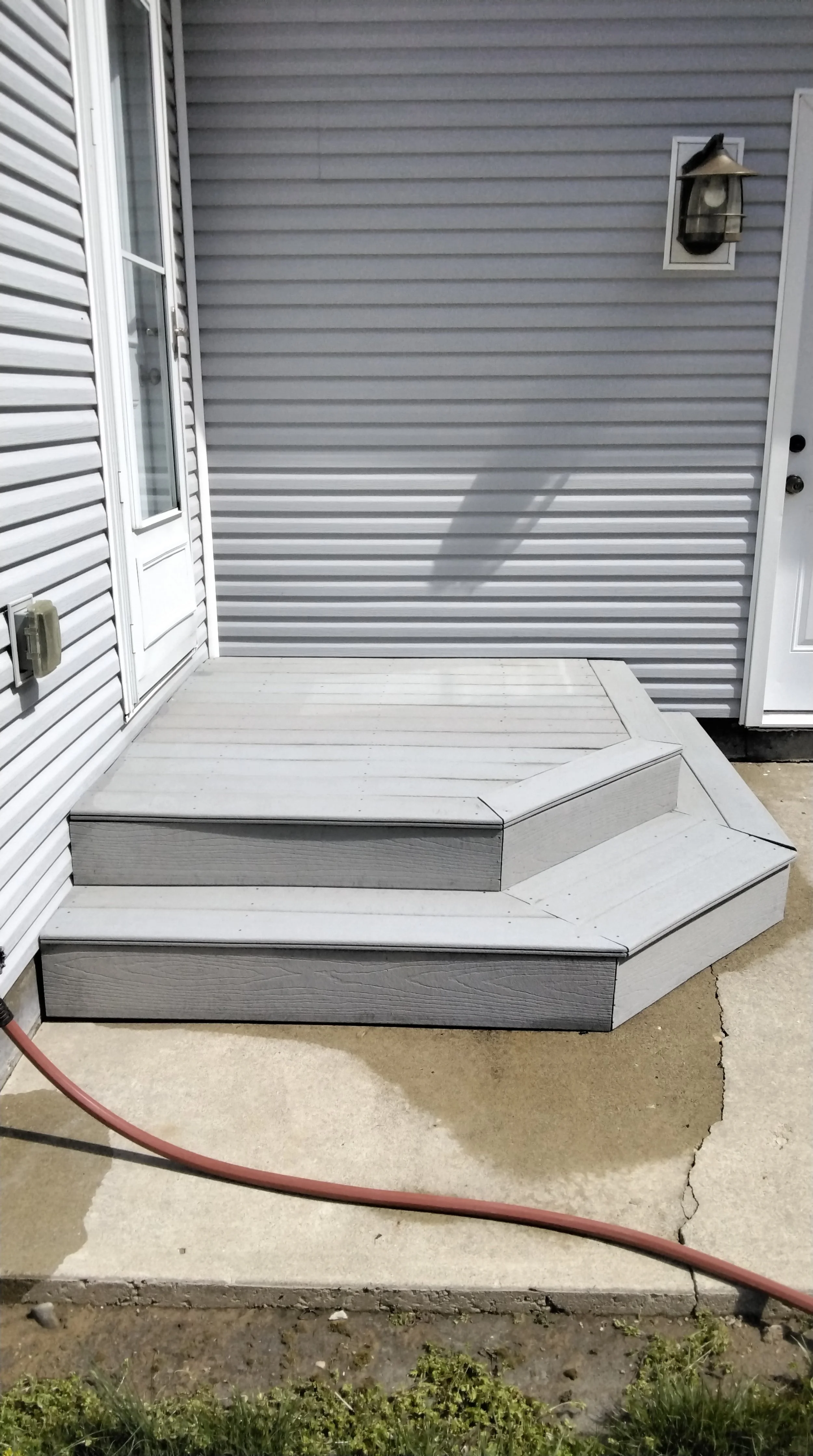 A small set of grey wooden steps leading up to a porch with concrete underneath, beside a white vinyl side wall of a house with a storm door and an exterior light fixture.
