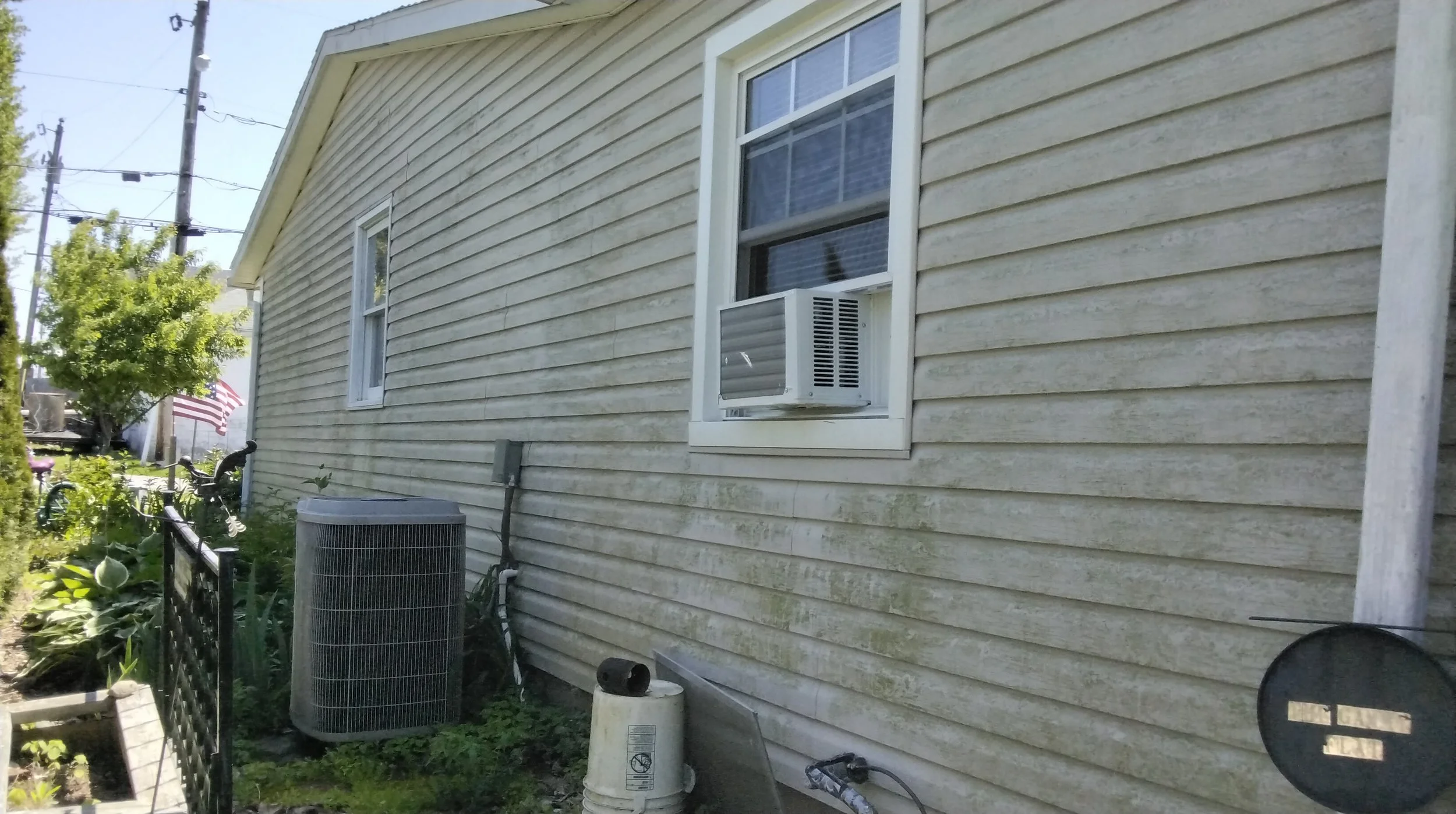 Side view of a house with beige vinyl siding, a window with an air conditioning unit, and a small garden with plants, a black fence, and outdoor equipment including an air conditioning compressor.