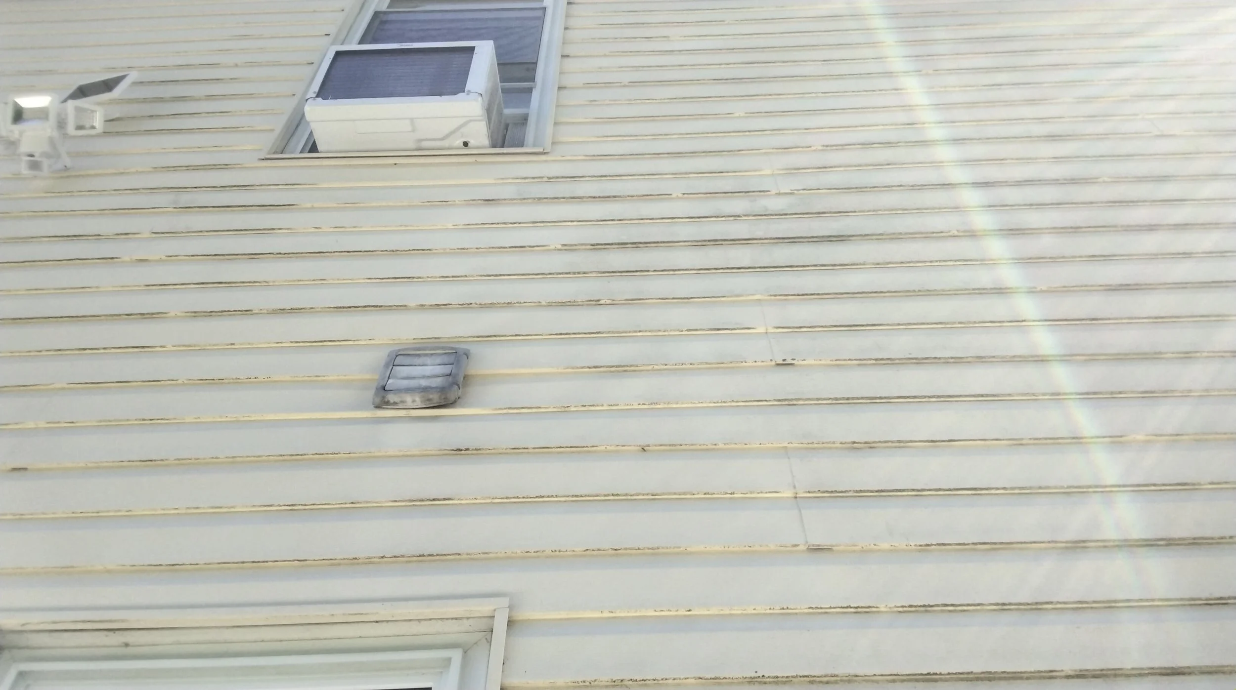 Close-up view of a house's beige siding exterior, showing a vent, a window with a handle, and an air conditioning unit in the window, with sunlight and a lens flare.
