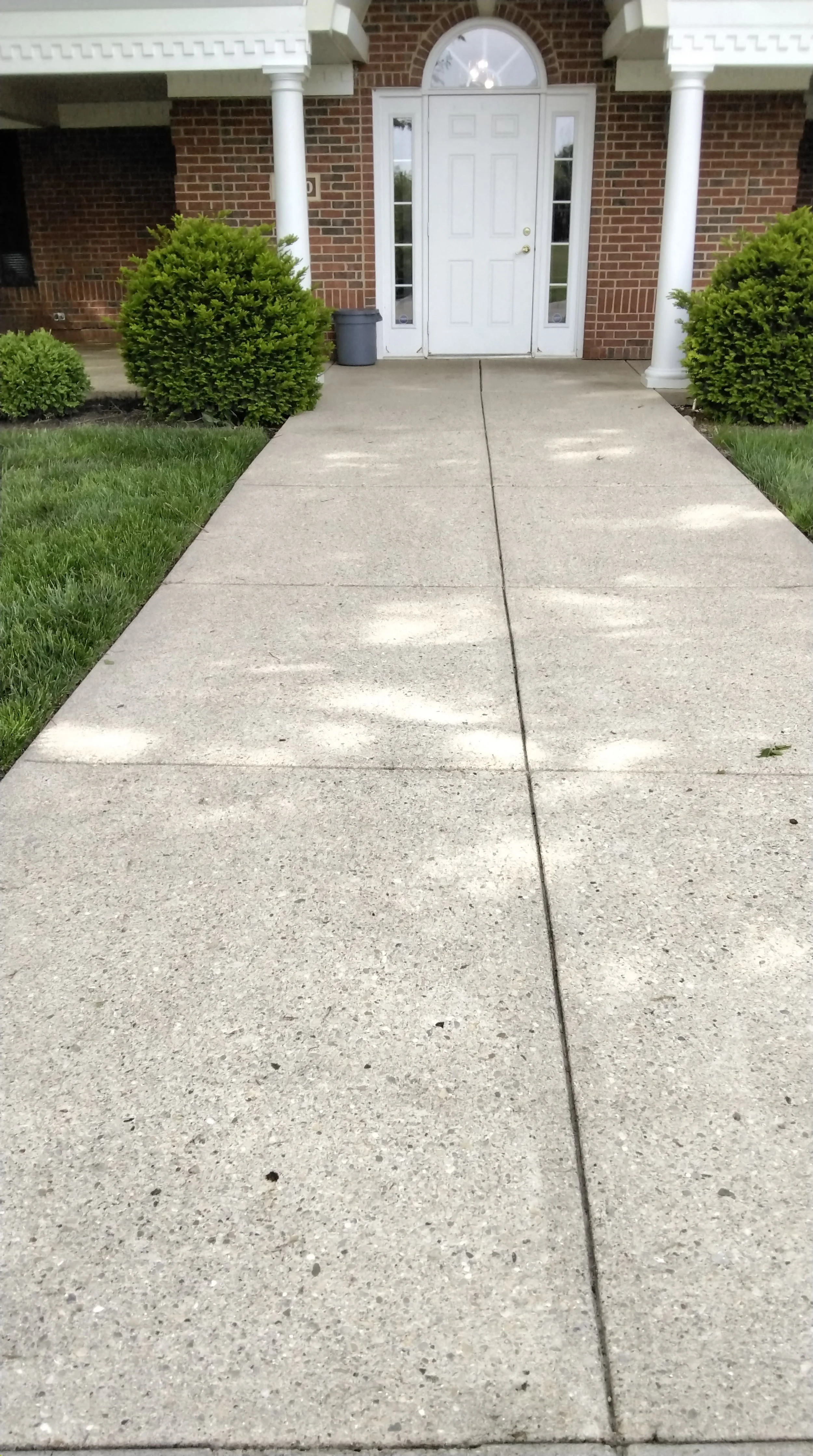 Concrete walkway leading up to a white front door of a brick house, flanked by bushes and supported by white columns.