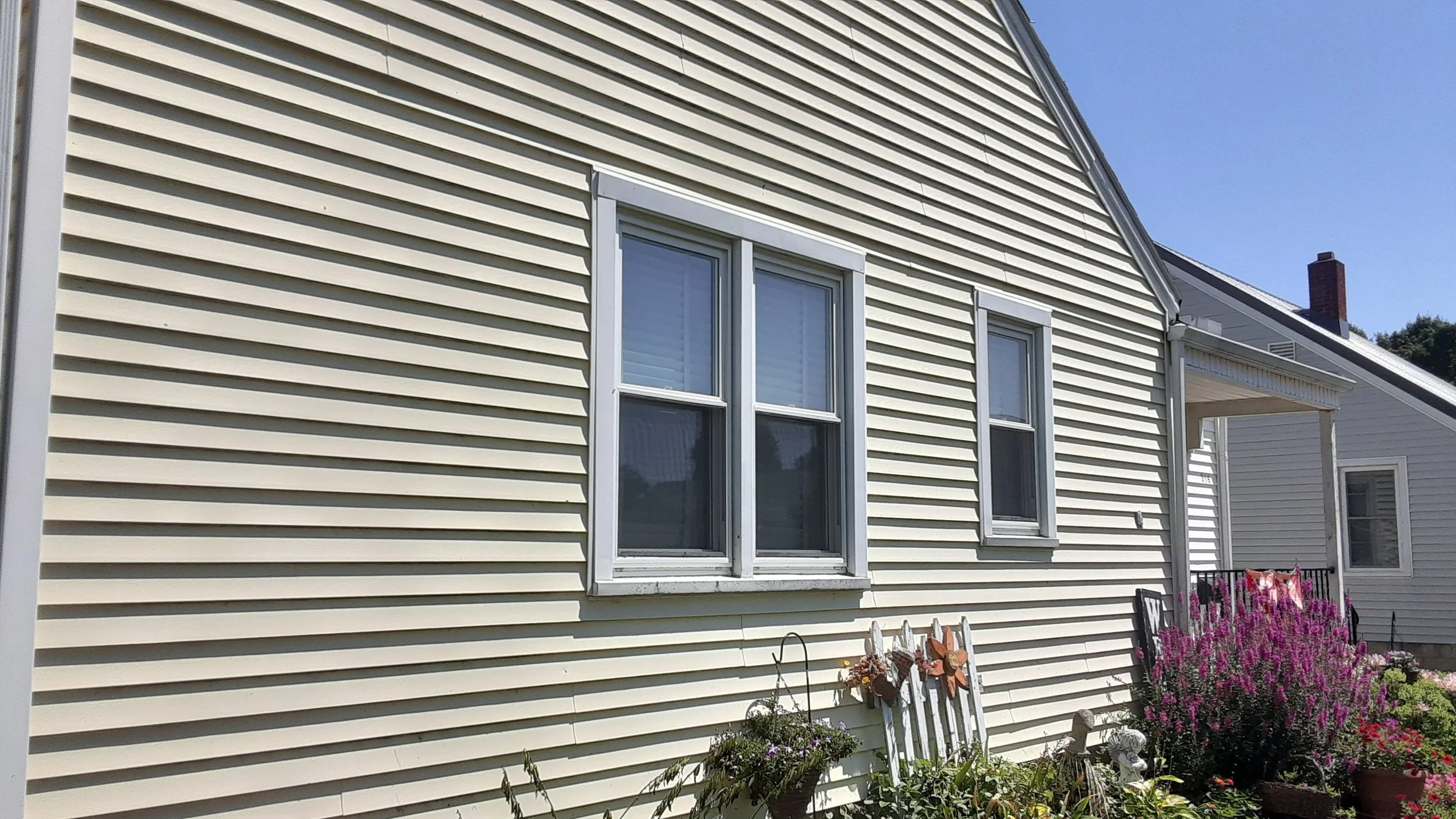 A house with beige vinyl siding, three double-hung windows with white frames, a small covered porch, and a garden with pink flowers and decorative items.