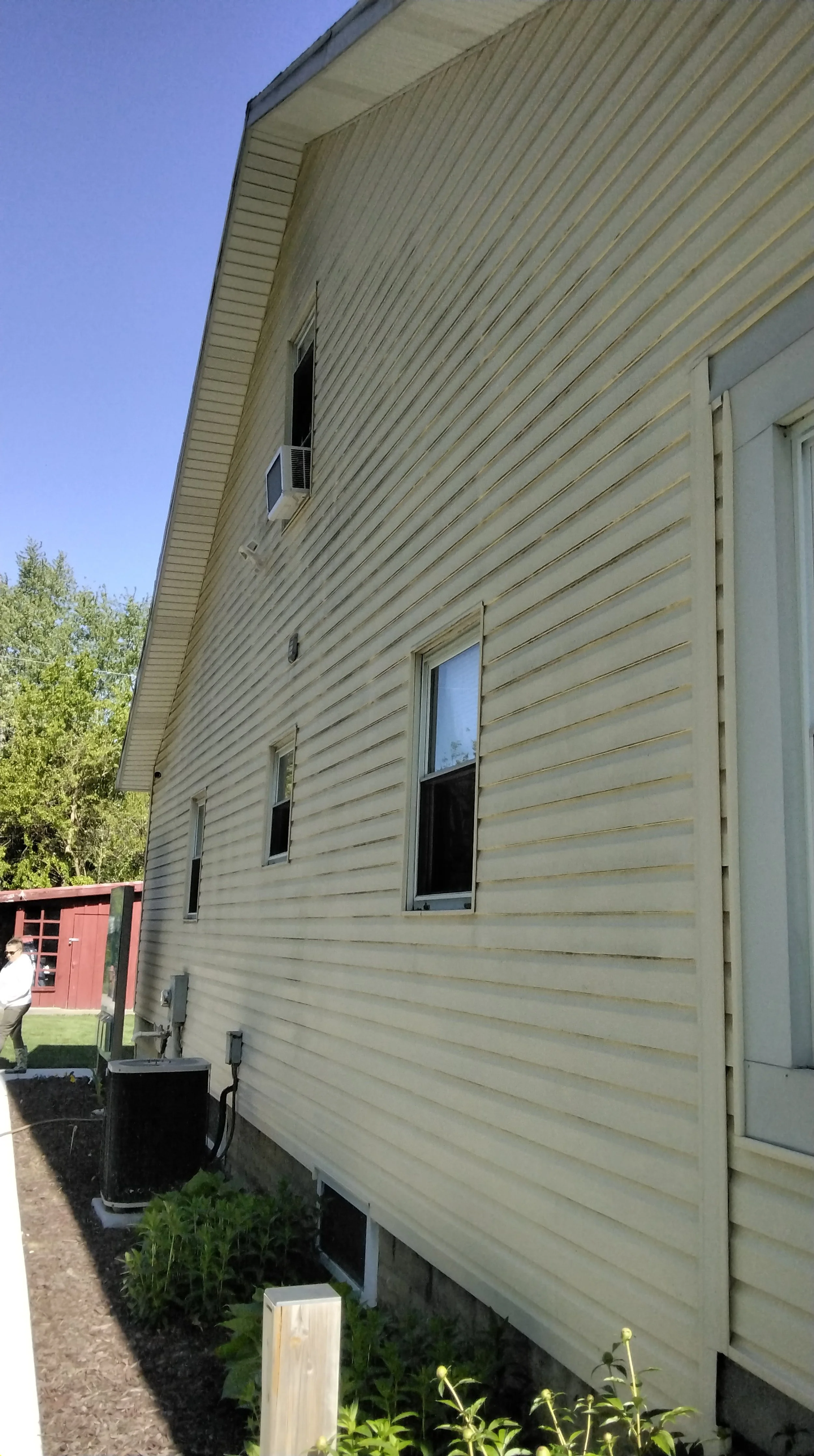 Side view of a beige house with several windows, an air conditioning unit, and vegetation at the base, with a person walking in the background near a red shed and a tree.
