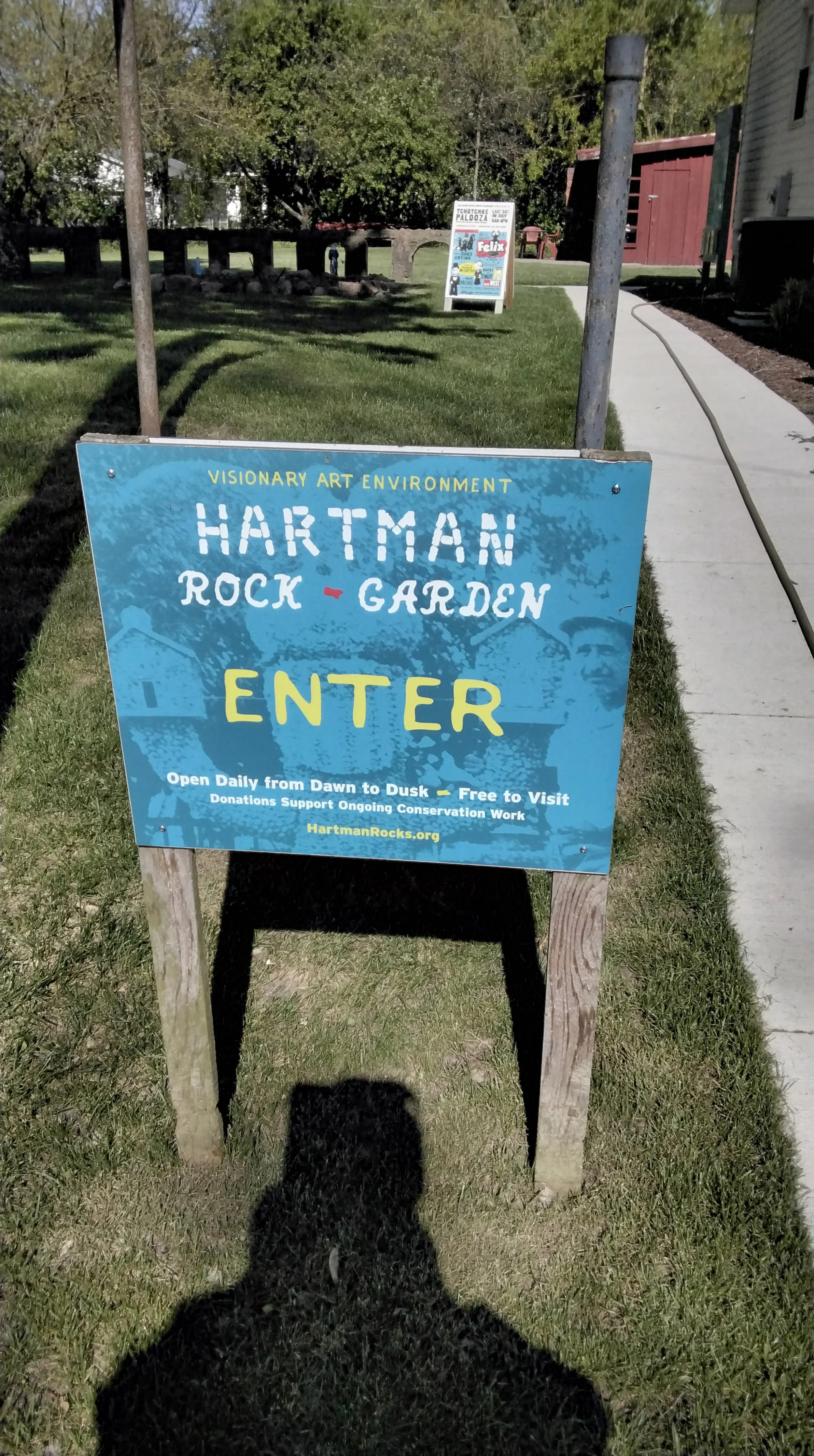 Blue signboard for Hartman Rock Garden with details about open hours, free admission, and conservation support, situated on a grassy area near a sidewalk with trees in the background.