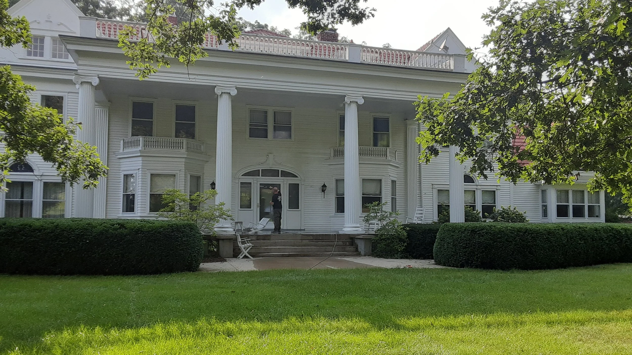 A large, white, two-story house with tall columns and a porch, surrounded by green grass and trees, with a person standing on the steps.