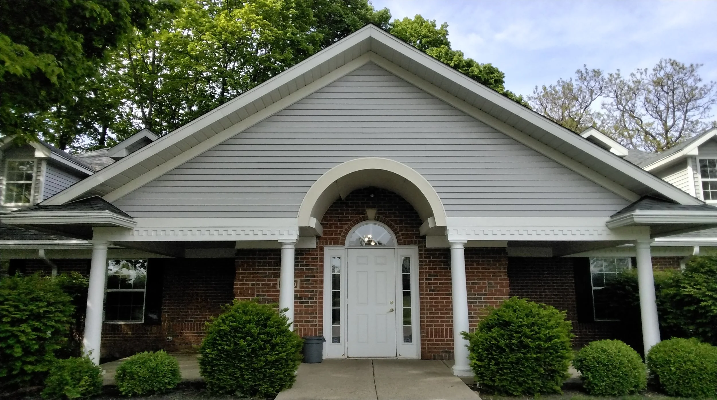 Exterior view of a house with a white front door, arched entryway, brick and white siding, surrounded by neatly trimmed bushes, with trees and a partly cloudy sky in the background.
