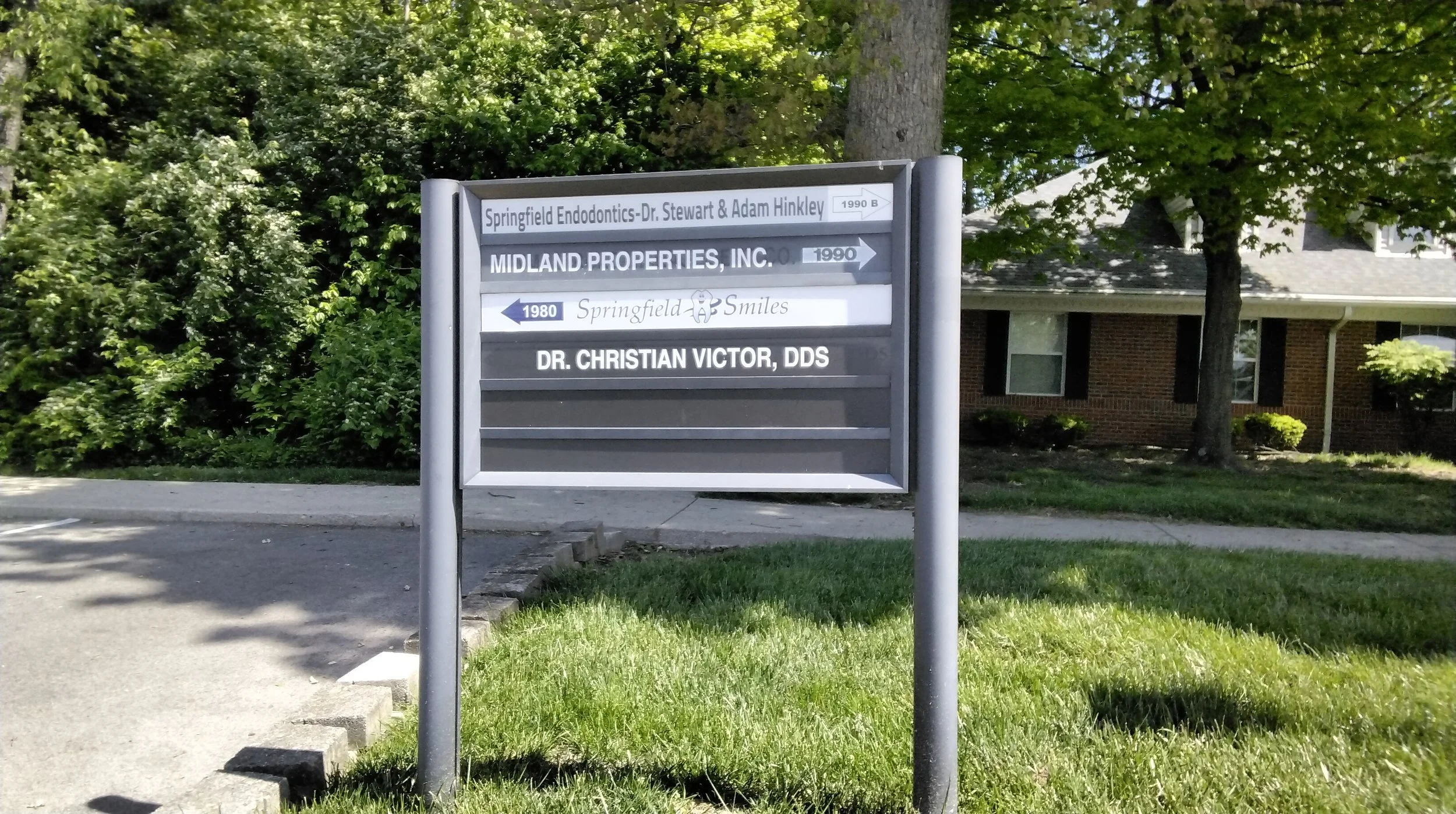 Signboard outside buildings with trees and a house in the background, displaying business names and dates, including Springfield Endodontics, Midland Properties, Springfield Smiles, and Dr. Christian Victor, DDS.