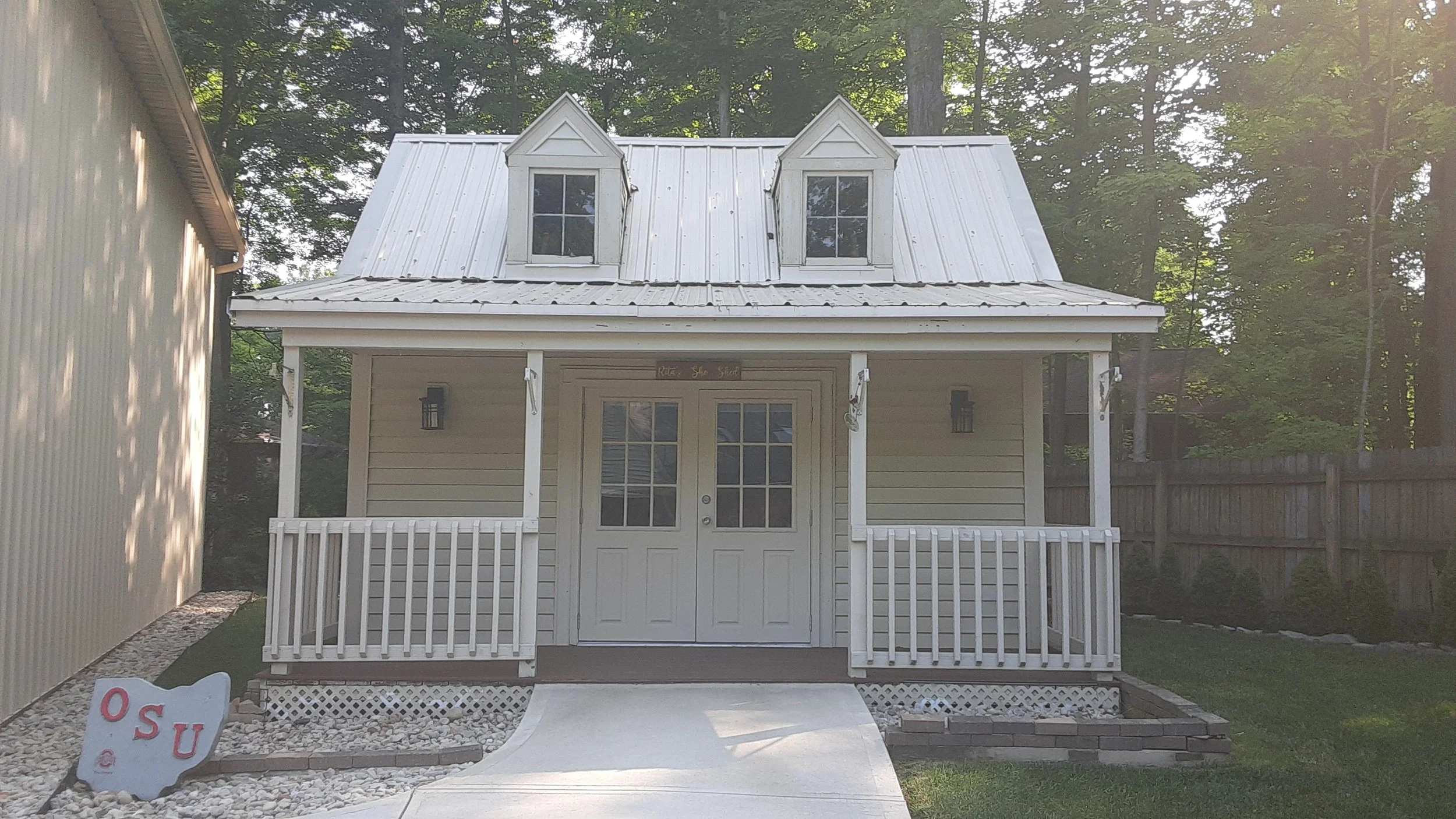 Small white house with two dormer windows, white metal roof, small front porch with white railing, and a concrete ramp leading to the door. There is an Oregon State University sign on the lawn in front.