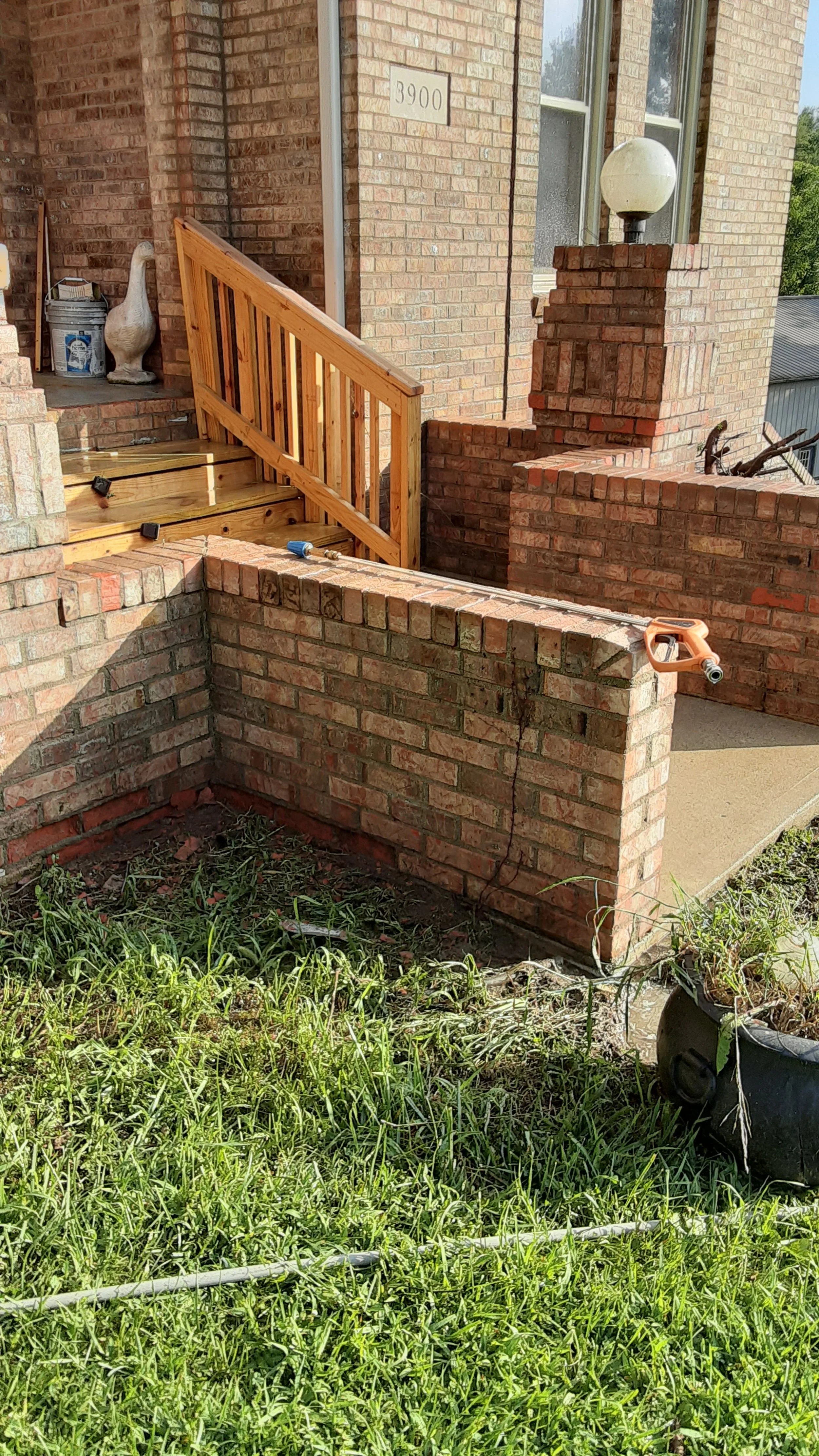 Brick house entryway with wooden porch steps and railing under construction, with stair tools and a garden. The house number 3900 is visible on the brick wall.