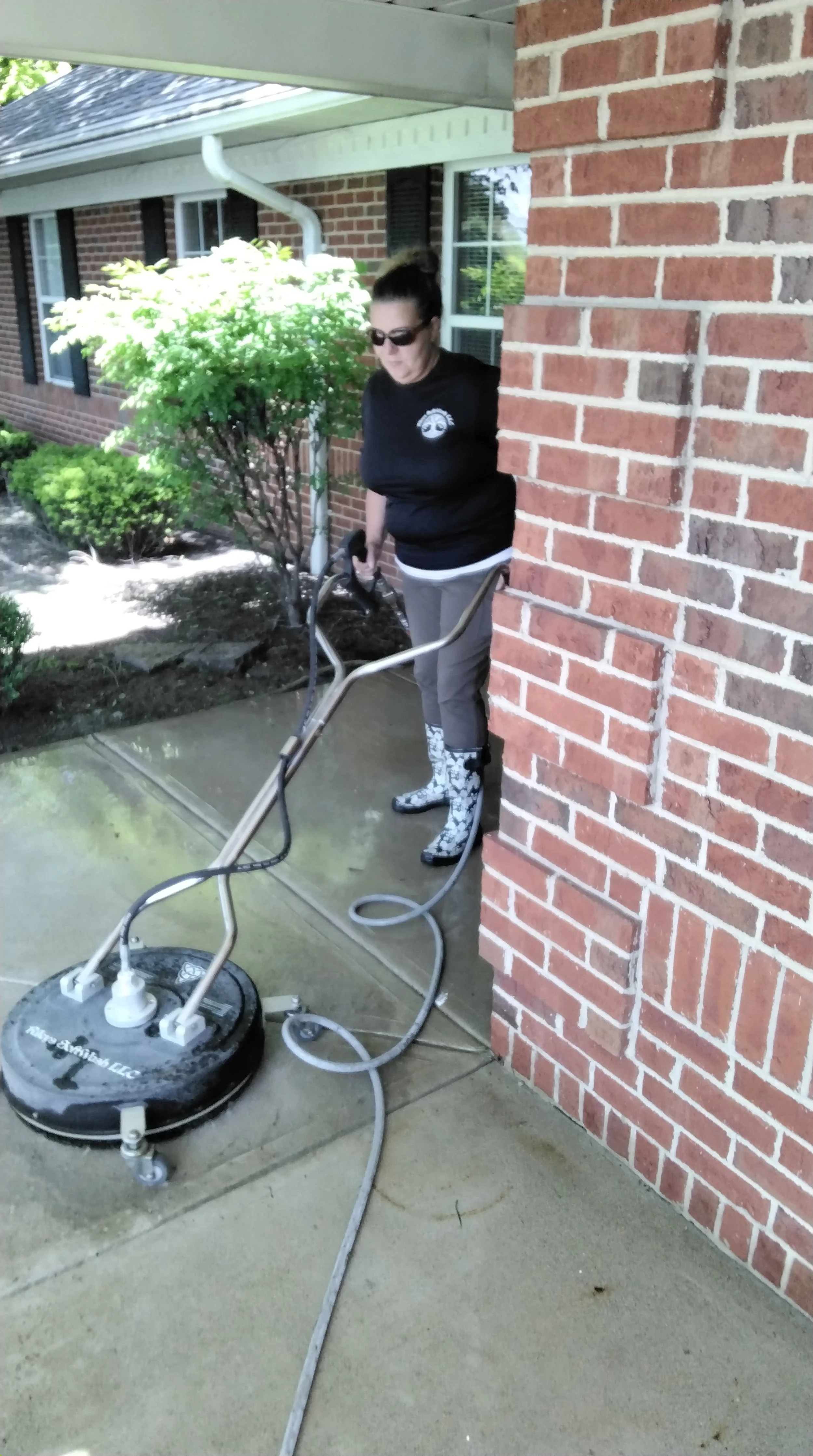 Woman wearing sunglasses and rain boots operating a surface cleaner on a concrete sidewalk beside a brick house.