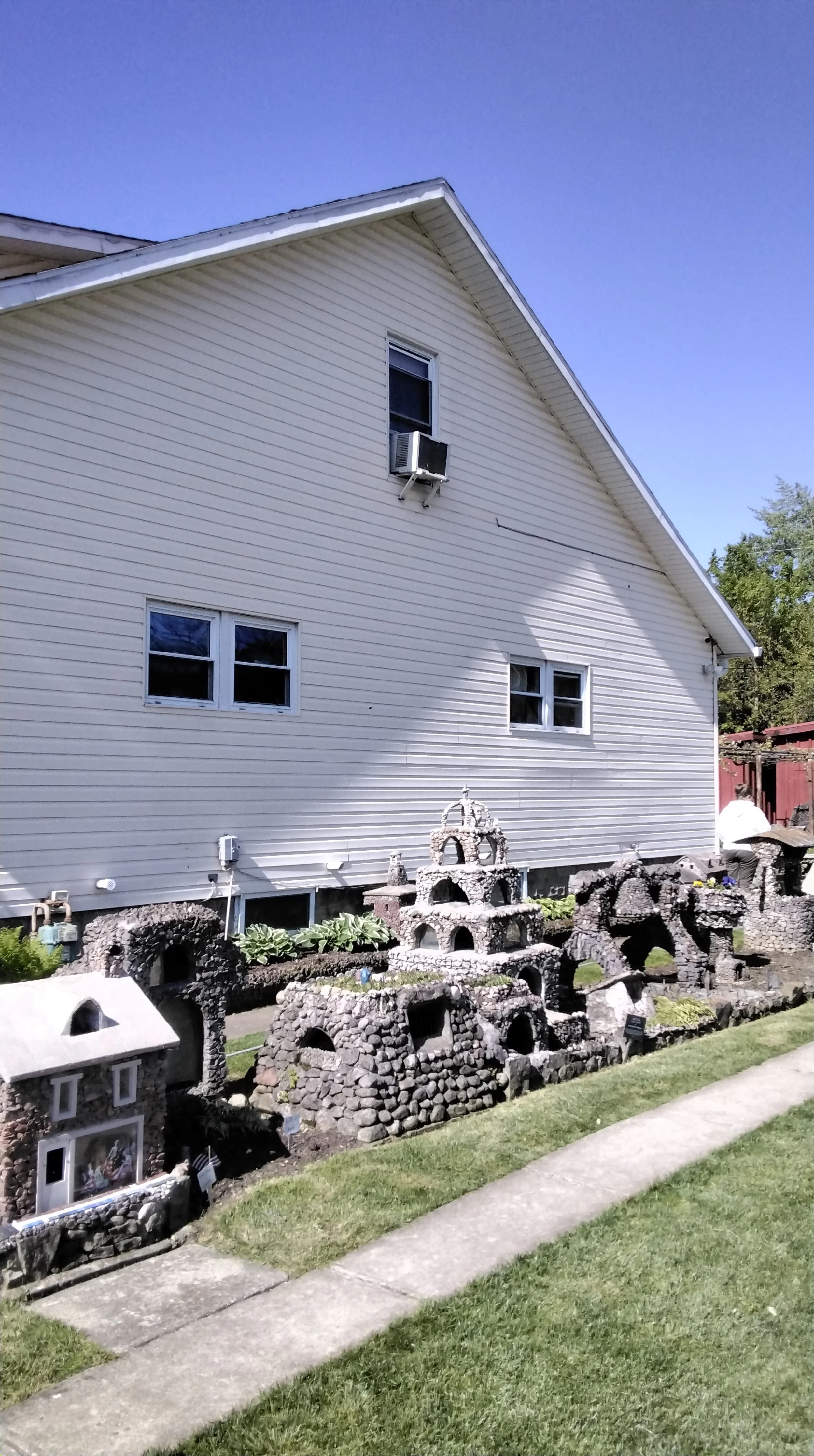 A white house with horizontal siding, three windows on the side wall, and an air conditioning unit in the upper window. In front of the house, there is a garden with small decorative stone structures and miniature houses made from rocks and mortar, w
