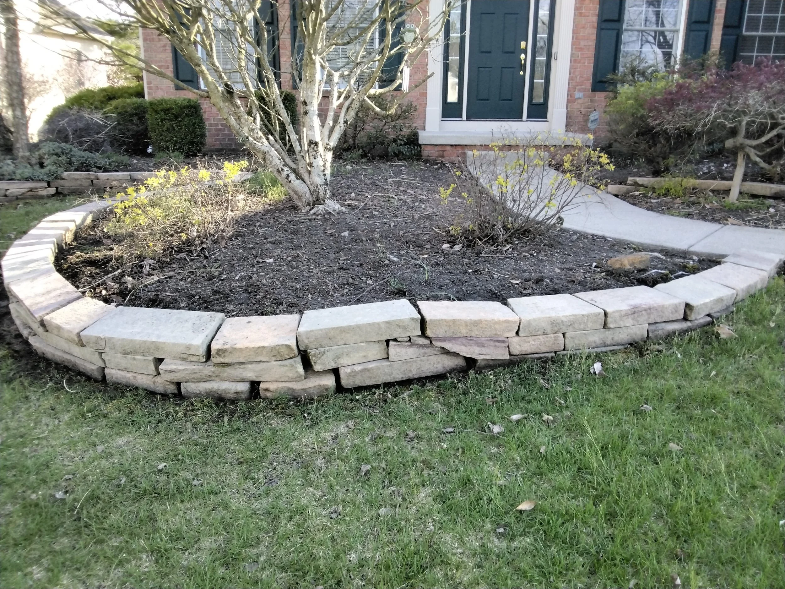 Front yard with a raised flower bed made of stacked stone bricks, containing a small tree and some bushes, with the entrance to a house with a dark green door in the background.