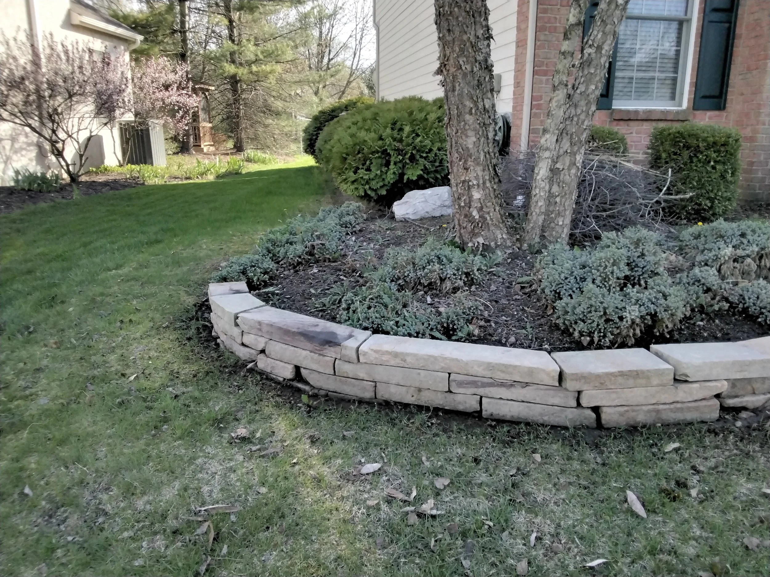 A landscaped yard with a curved stone retaining wall, green grass, blooming trees, and shrubs near a house with brick and siding exterior.