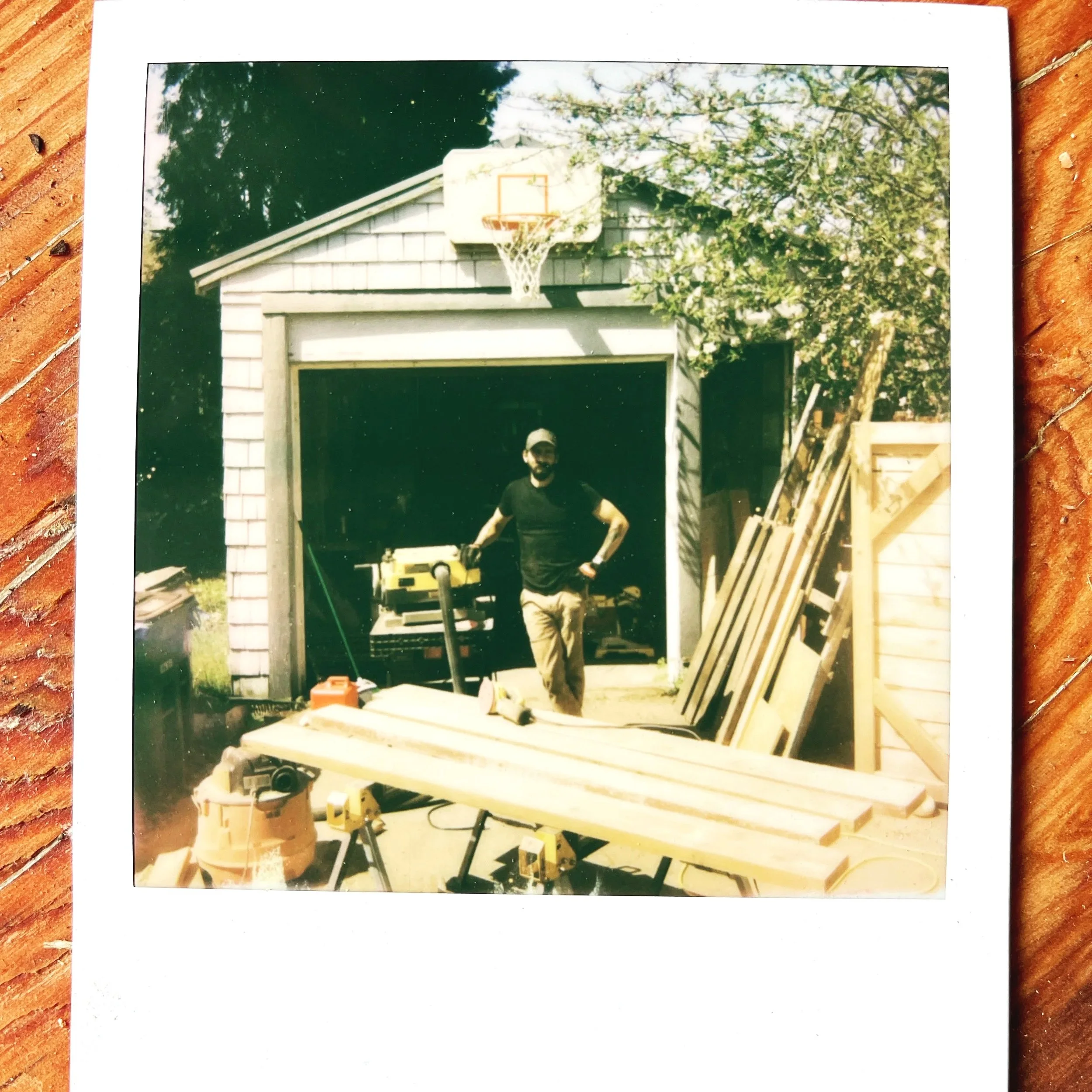 A person standing in a garage during construction, surrounded by wood planks, with a ladder, power tools, and a partially built structure.