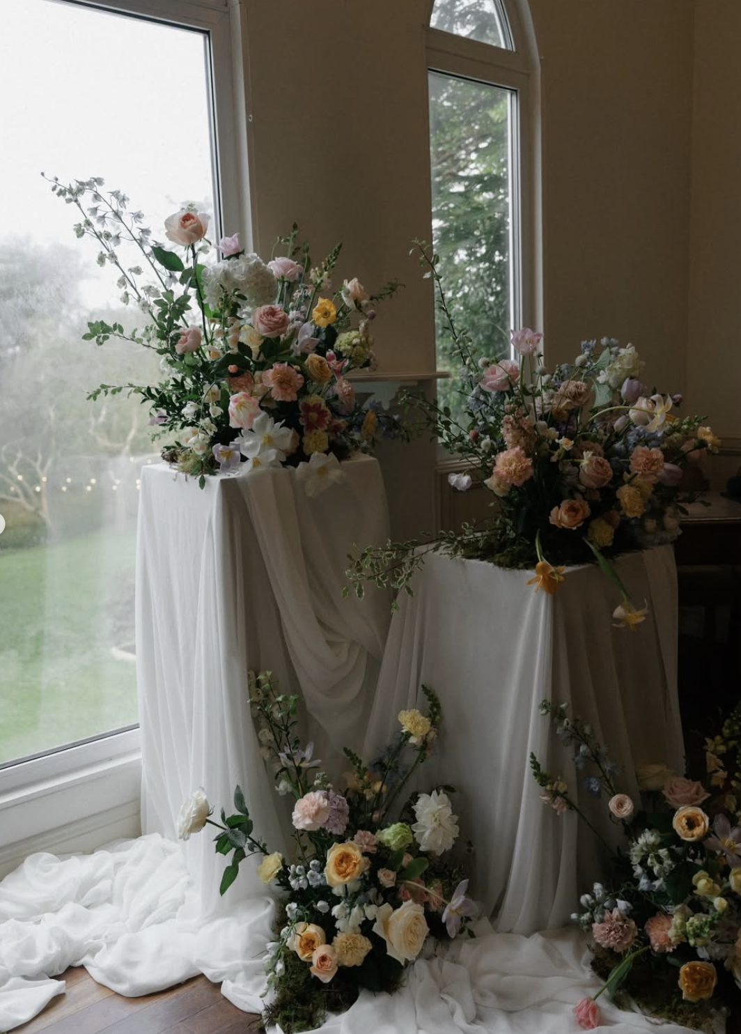 Elegant floral displays with pastel roses, peonies, and greenery on white draped fabric near large windows.