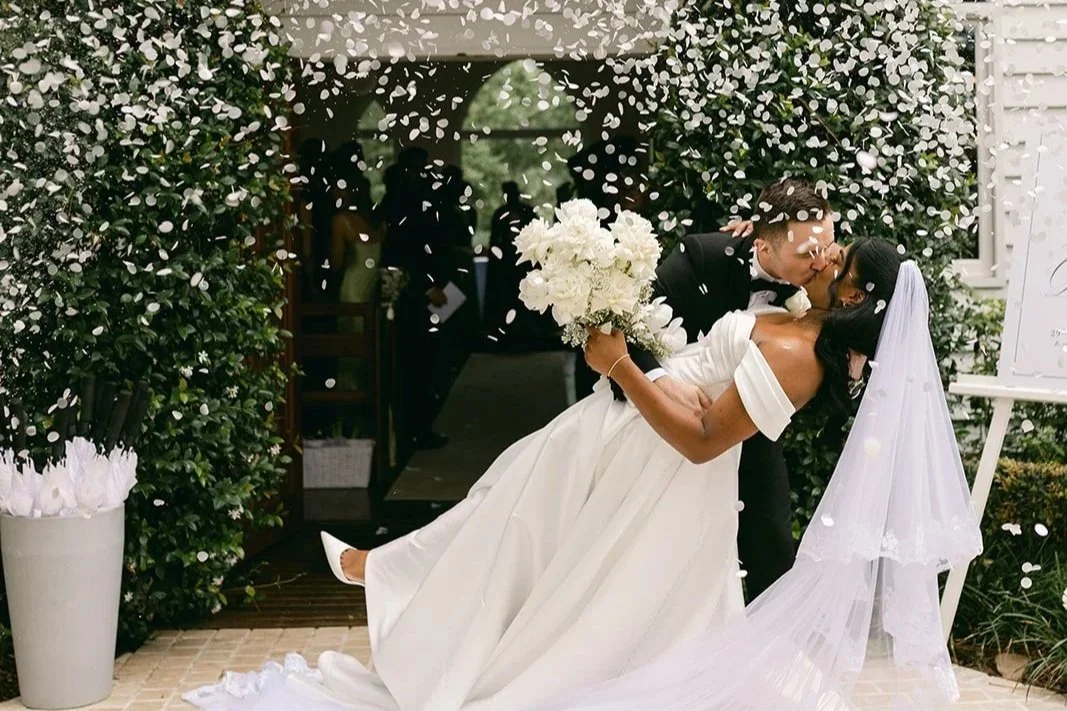 A newlywed couple sharing a kiss during a wedding celebration, with the groom dipping the bride, who is holding a bouquet of white flowers, surrounded by guests and confetti.