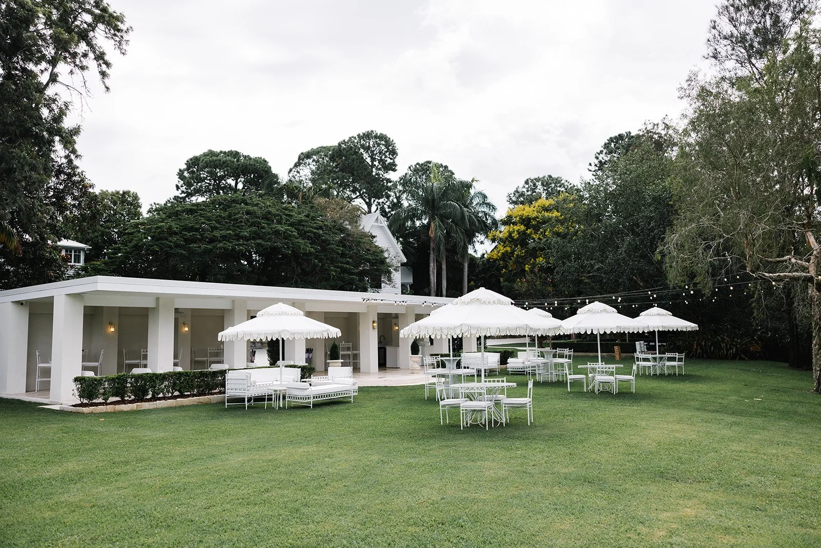 An outdoor event setup with white chairs, tables, and large white umbrellas on a grassy lawn, adjacent to a white building with a covered patio and string lights, surrounded by tall trees.