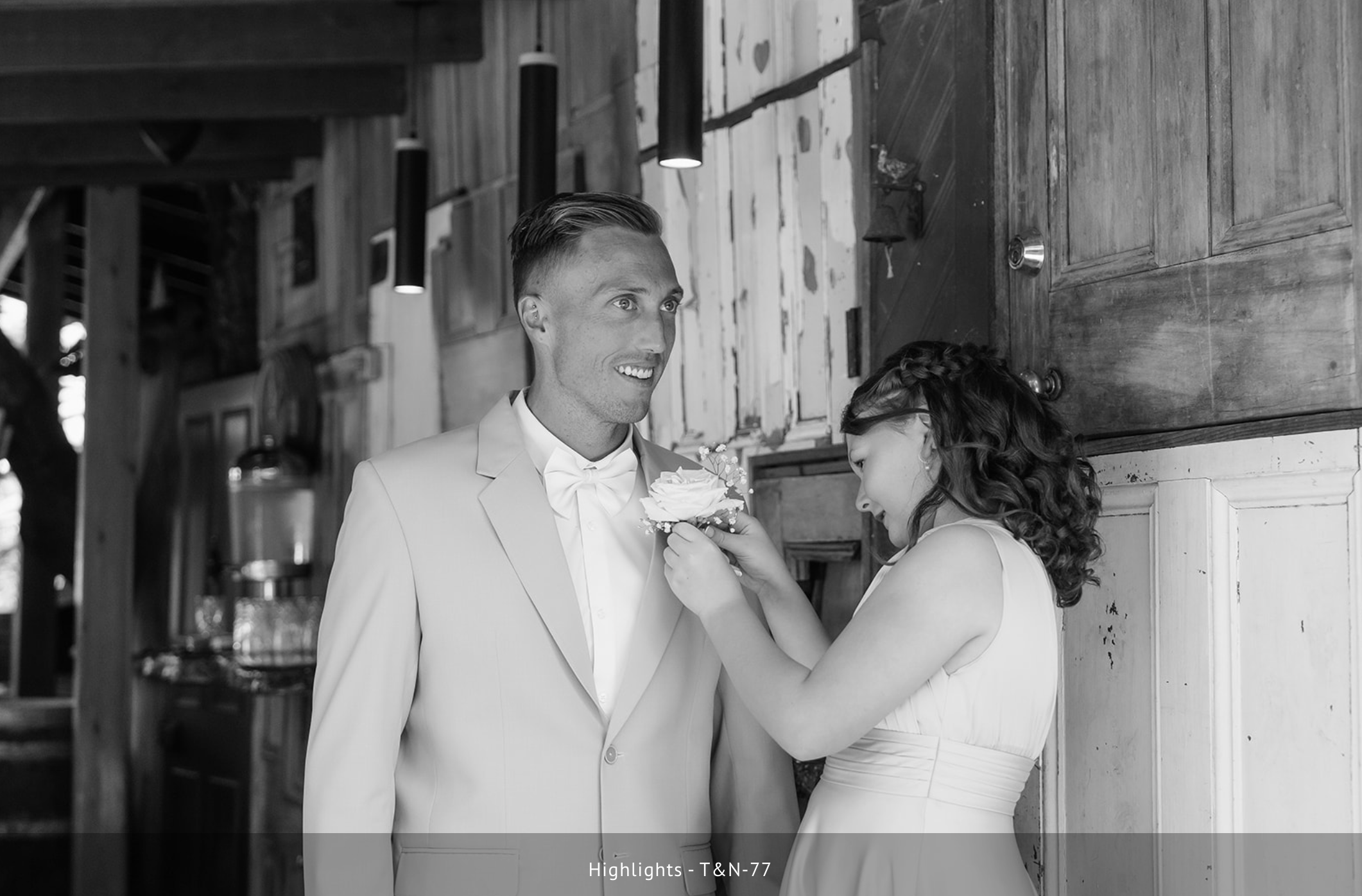A man in a tuxedo and a woman in a white dress sharing a moment indoors, the woman is holding a small bouquet and they are smiling.