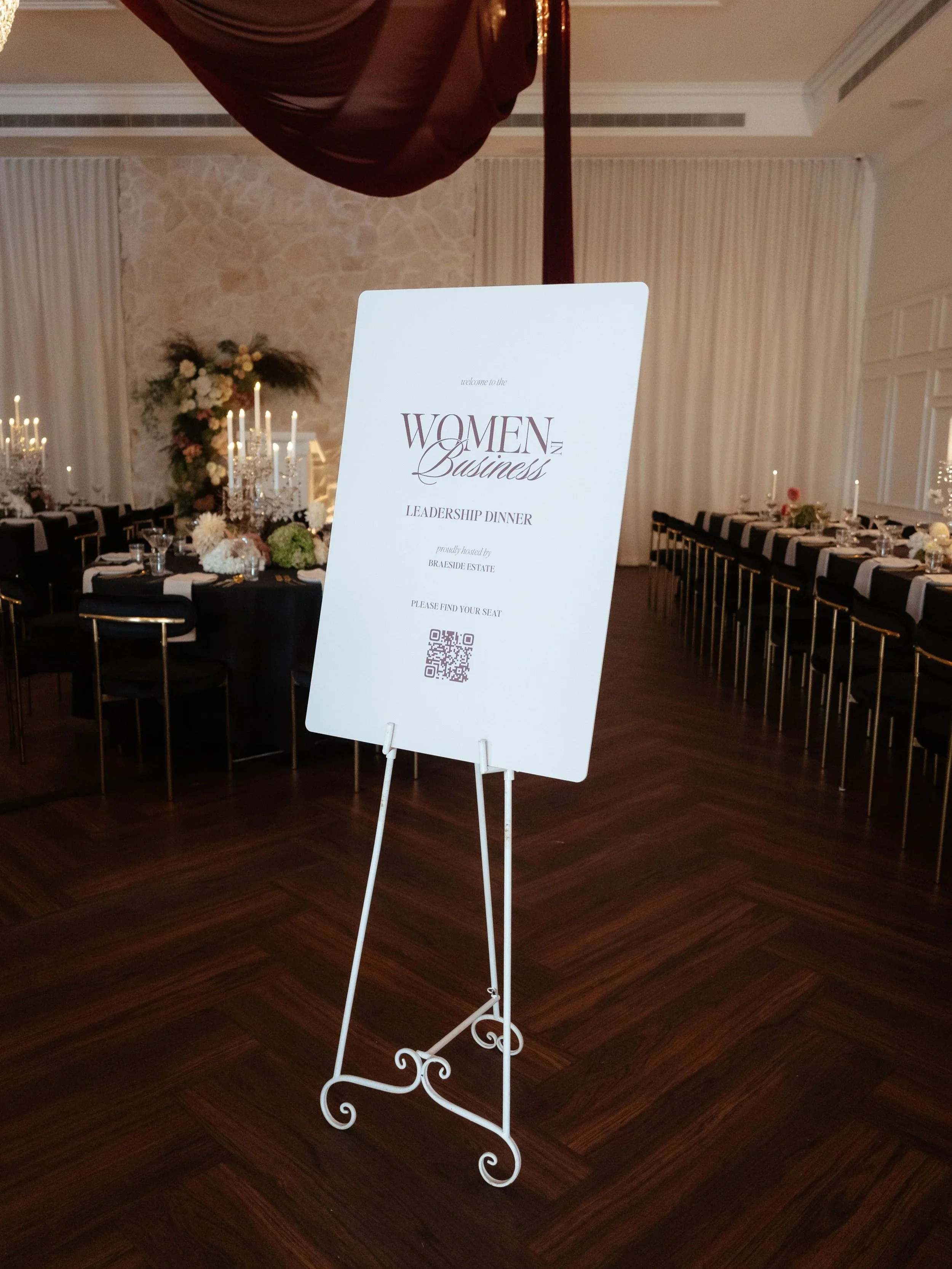 A white standing sign with event details in a decorated banquet hall. The sign reads 'Women in Business Leadership Dinner' hosted by Braeside Estate, with a QR code at the bottom. The background features round tables with black tablecloths, floral centerpieces, candles, and elegant place settings, and a floral arrangement and candles on a side table.