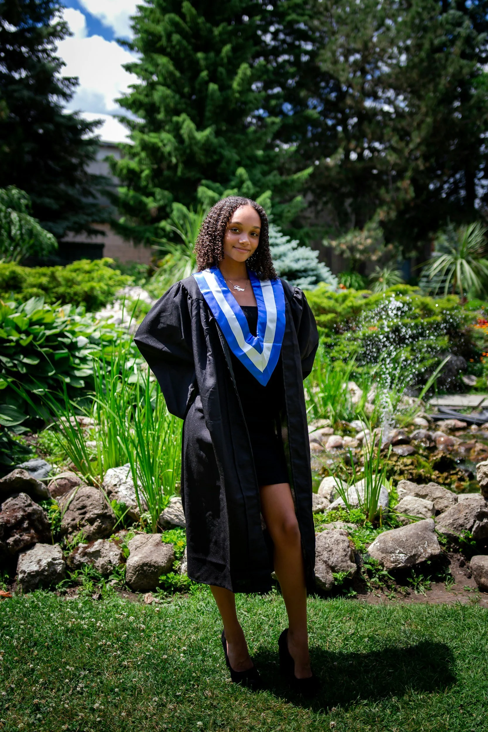 A young woman wearing a graduation gown and stole, standing outdoors in a lush garden with various plants, rocks, and tall trees in the background, smiling softly.
