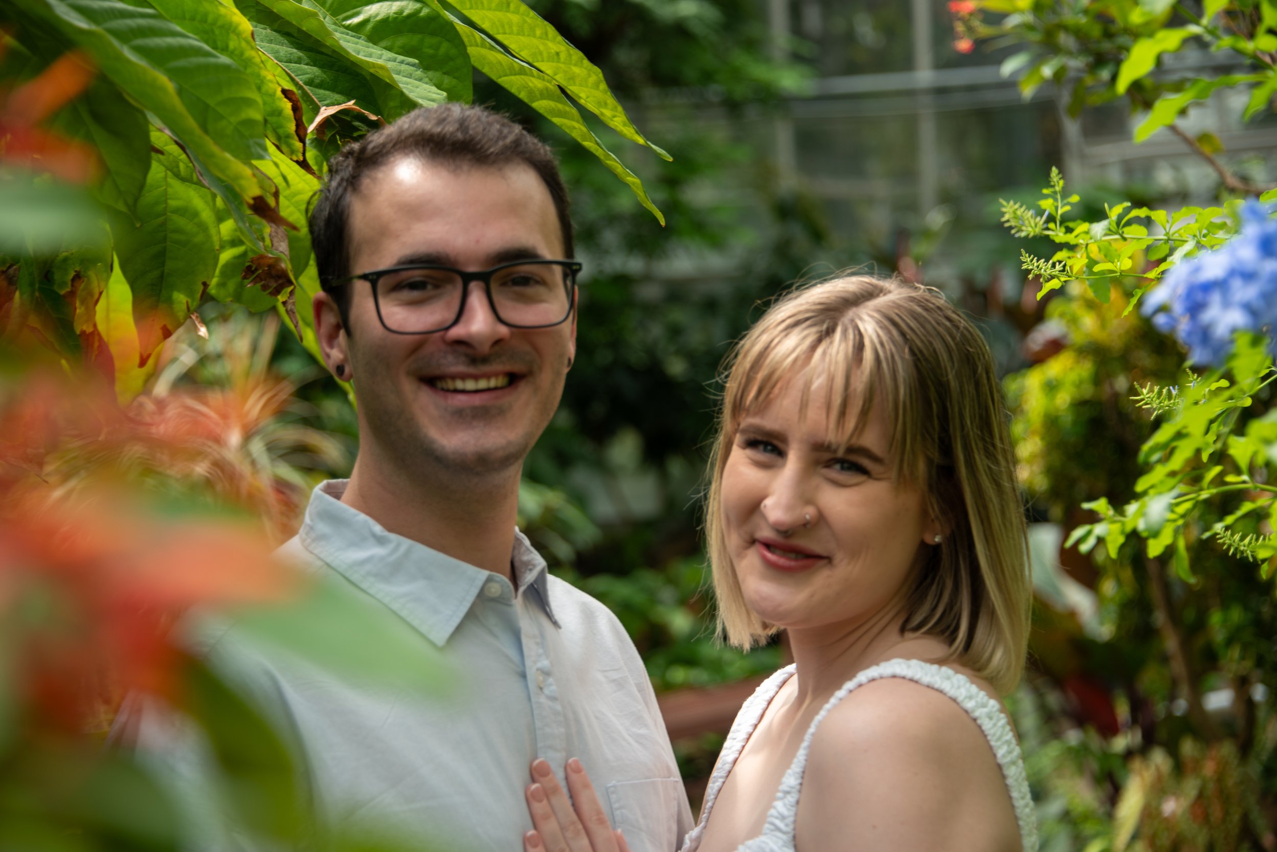 Smiling young man with glasses and short dark hair, and young woman with blonde hair and a nose piercing, standing close together outdoors surrounded by green foliage and flowers.
