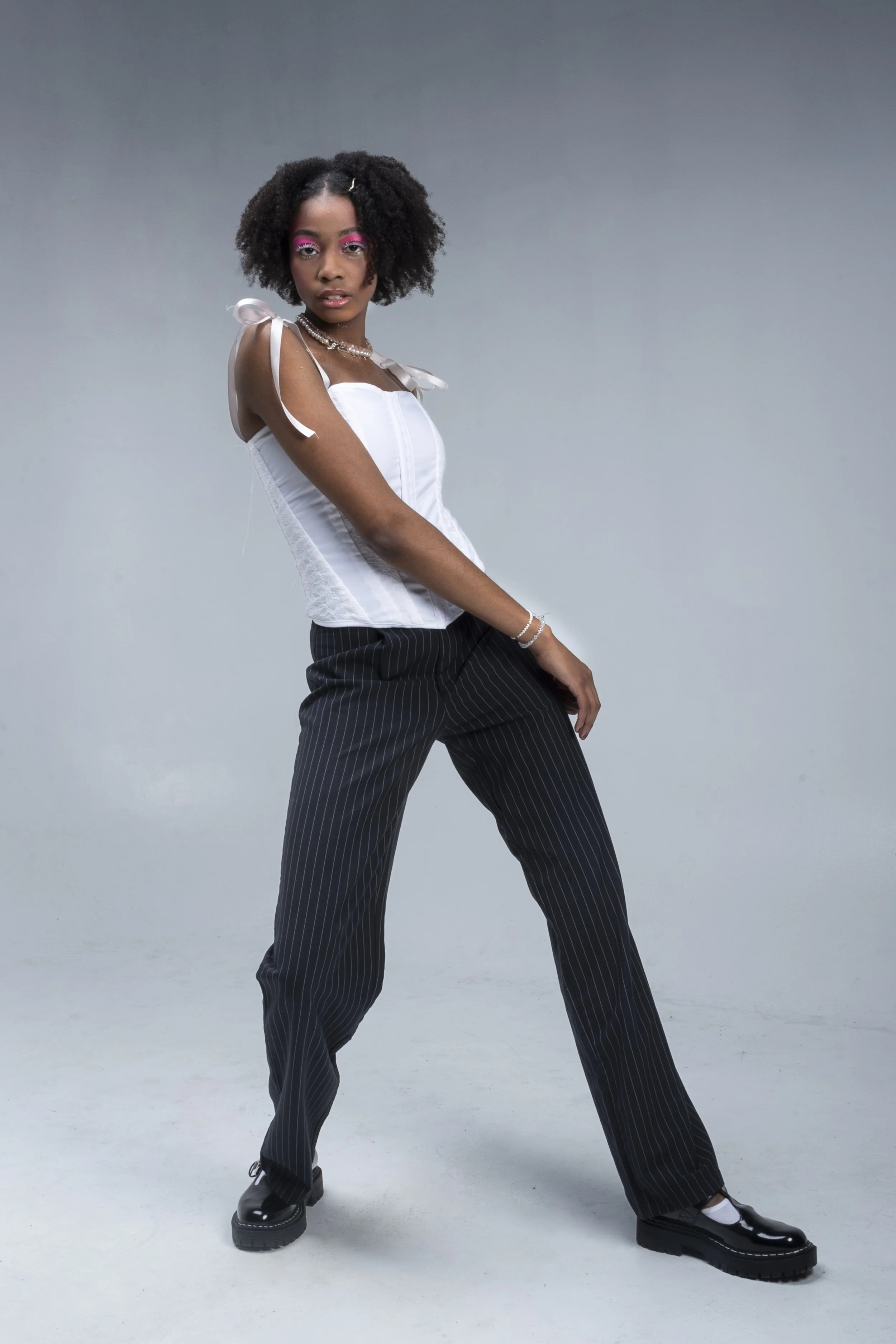 A young woman with curly hair and pink eye makeup wearing a white corset top with ribbon straps, striped black pants, and black platform shoes, standing against a plain grey background.