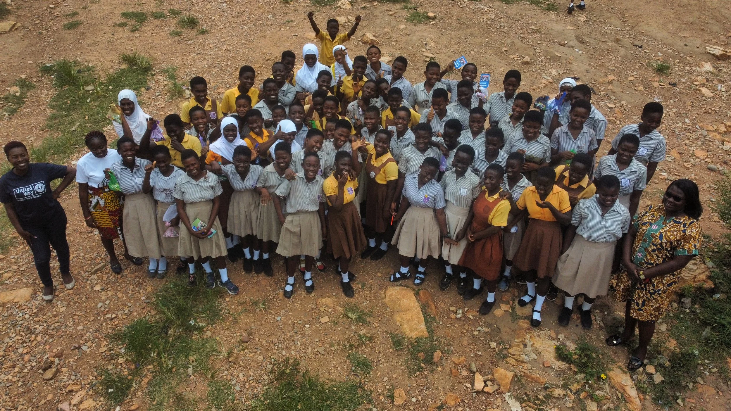 A large group of children and a few adults standing outdoors on rocky ground, smiling and waving at the camera, with some holding small items and banners.