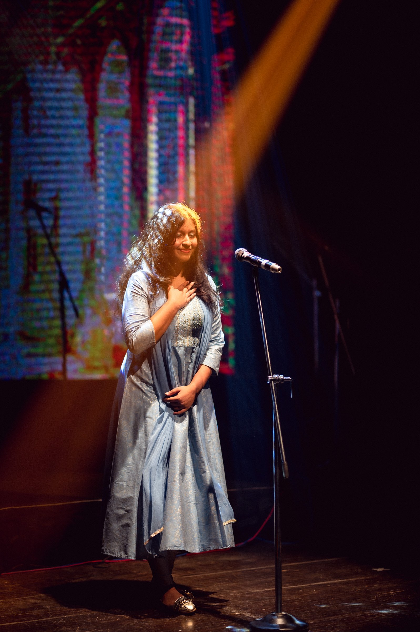 Person on stage holding a microphone, wearing a blue dress, with colorful background lighting.